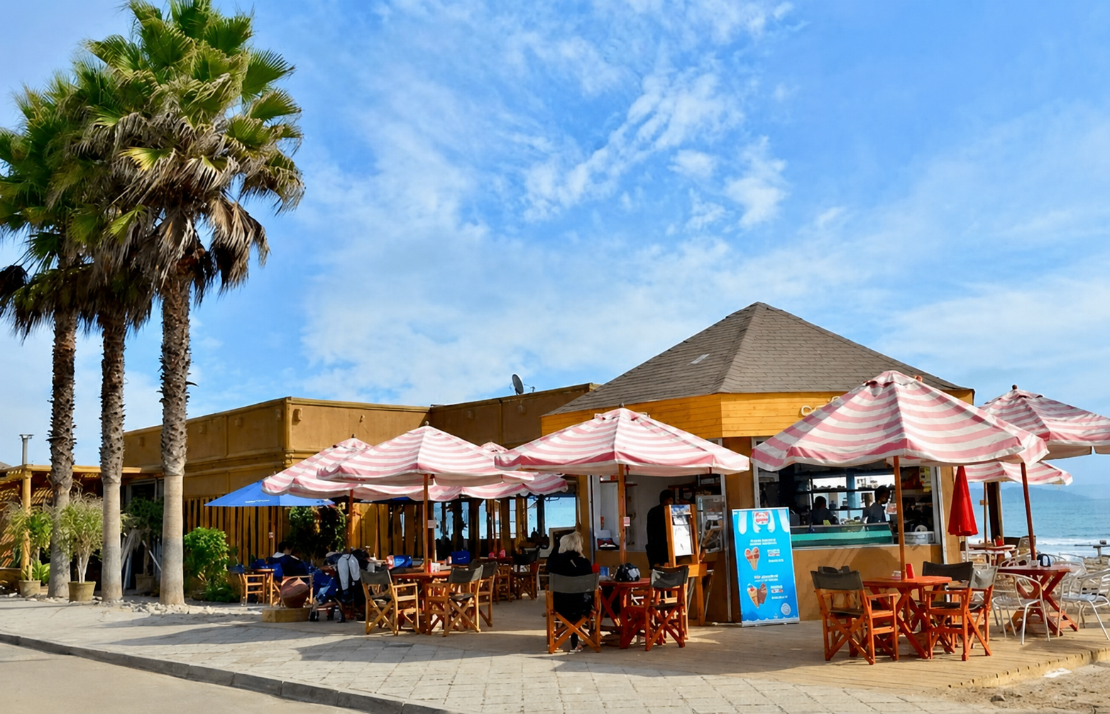 Restaurante en la playa con sombrillas rosas y blancas, palmeras y cielo azul con algunas nubes.
