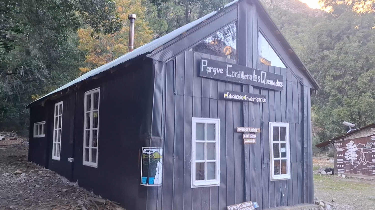 Una cabaña de madera negra con varias ventanas, localizada en un entorno natural con árboles y colinas. Hay un cartel en la cabaña que dice "Parque Cordillera Los Quemados" y otros signos relacionados con educación, investigación y servicios administrativos.
