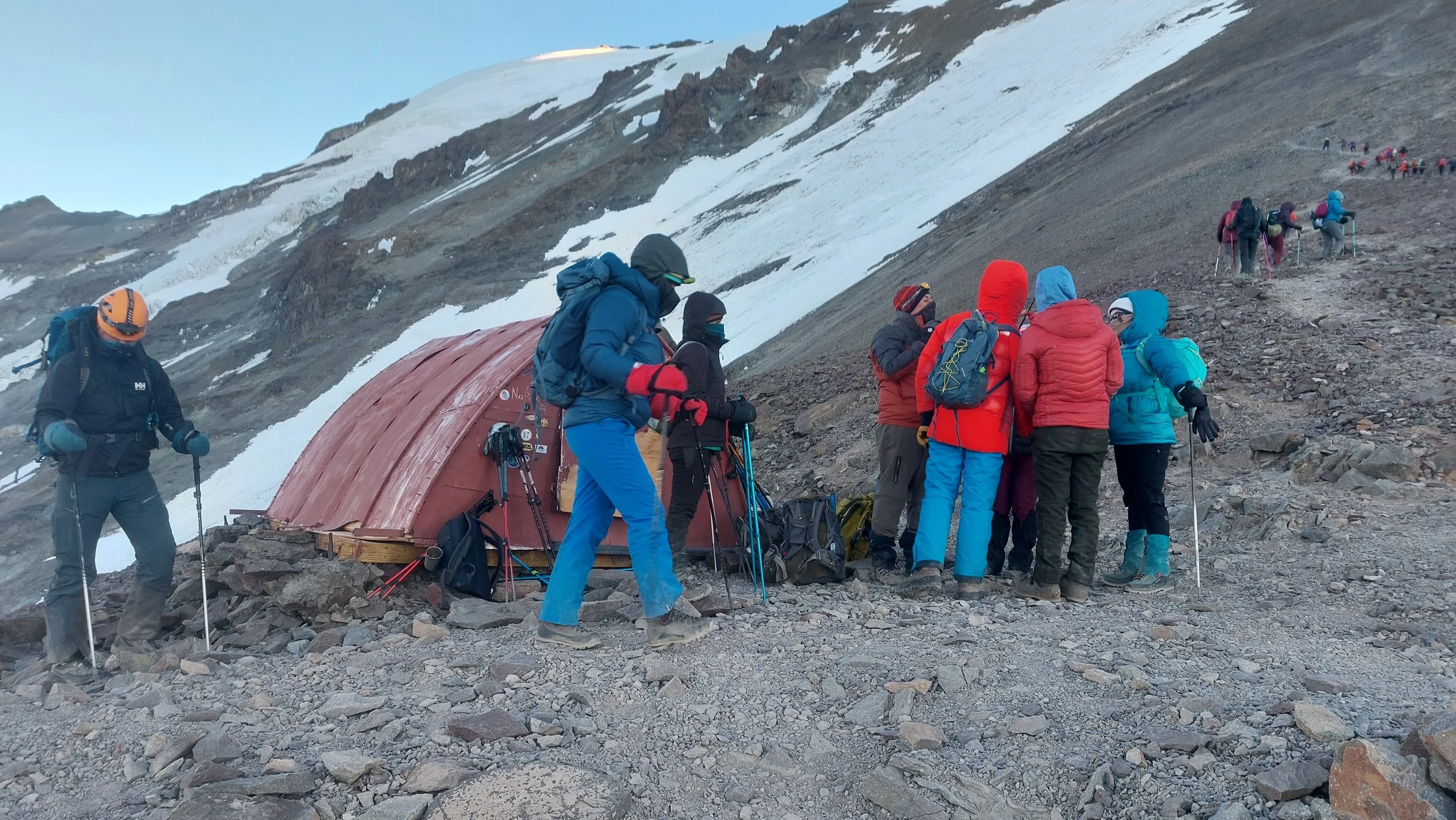 Grupo de personas haciendo senderismo en una zona montañosa con nieve, algunos descansando cerca de una estructura de refugio y otros subiendo en fila por el terreno rocoso.