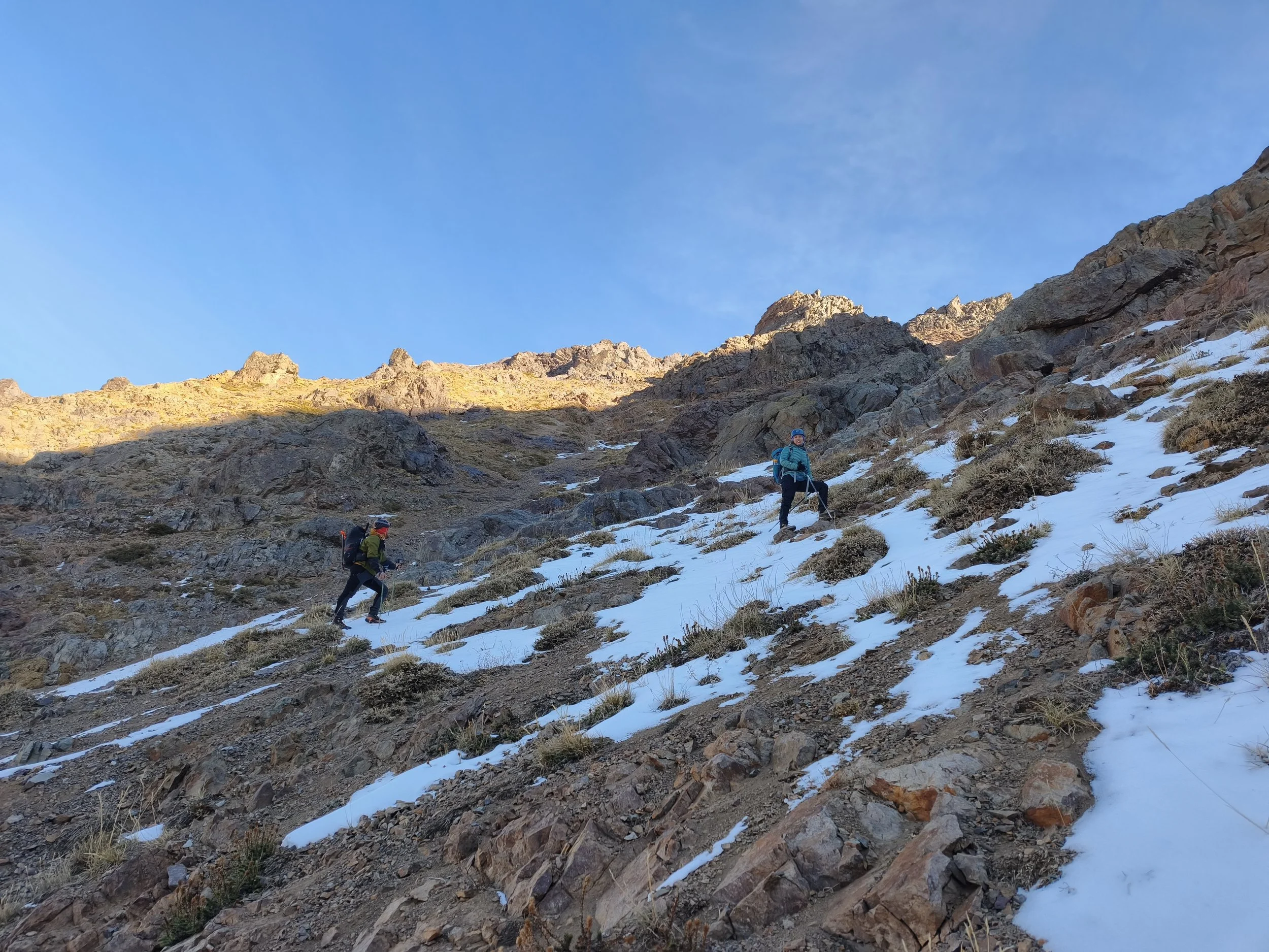 Dos personas de senderismo en montaña con nieve, rocas y un cielo azul despejado.