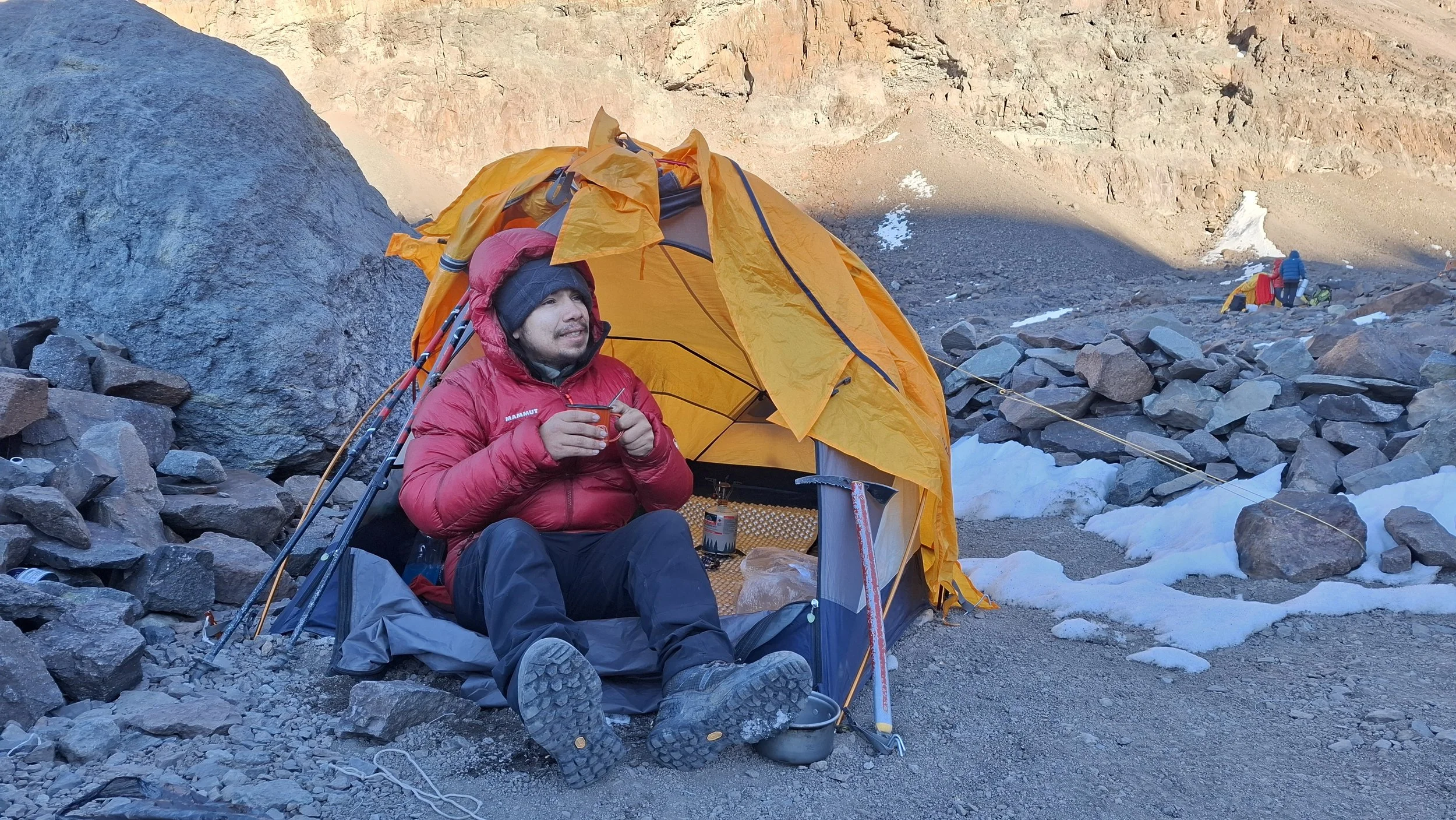 Persona descansando y tomando una bebida junto a una tienda de campaña naranja en un entorno de montaña con rocas y nieve.
