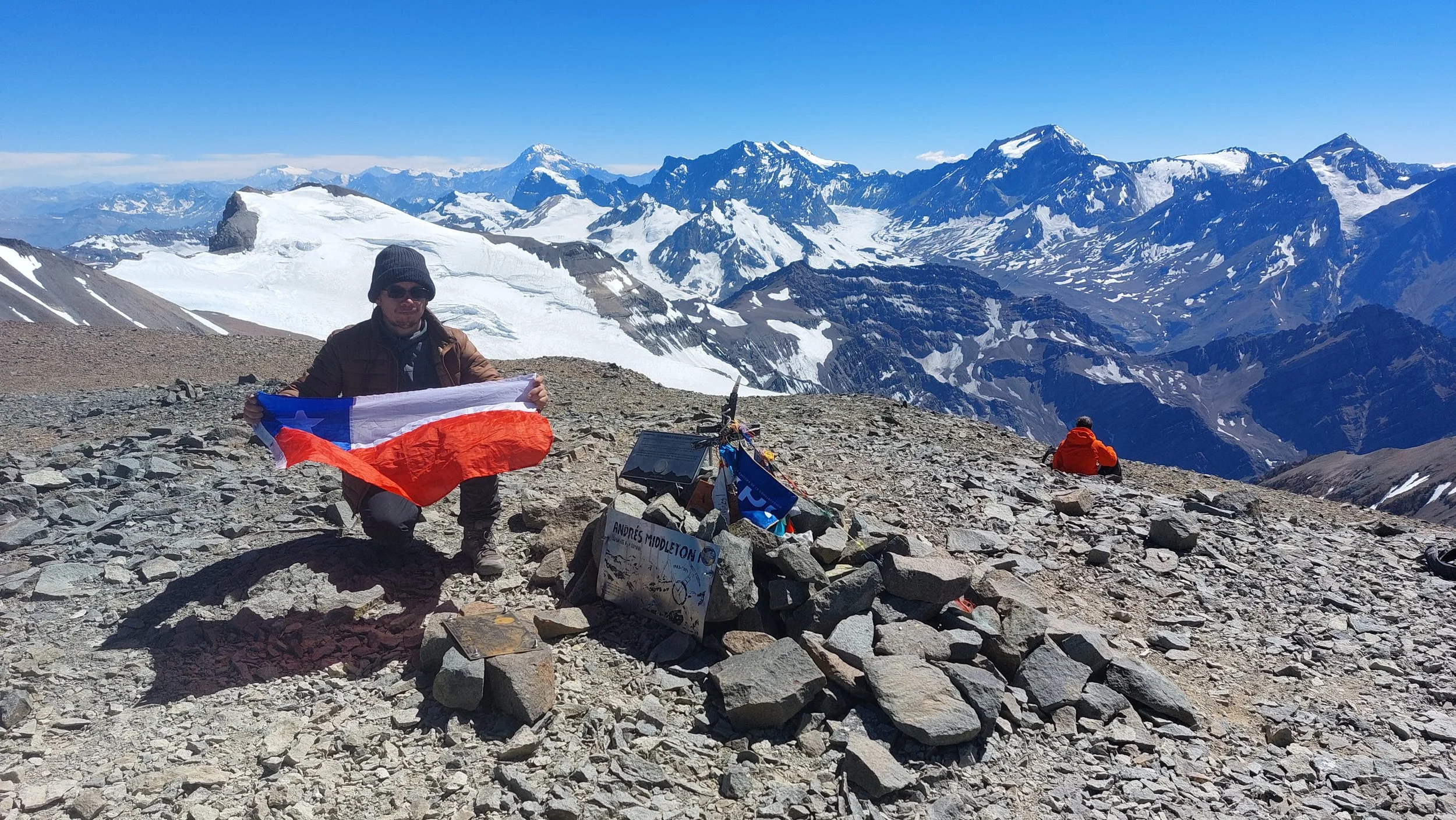 Persona en una cima montañosa, sosteniendo una bandera chilena, con picos nevados y un cielo despejado en el fondo.