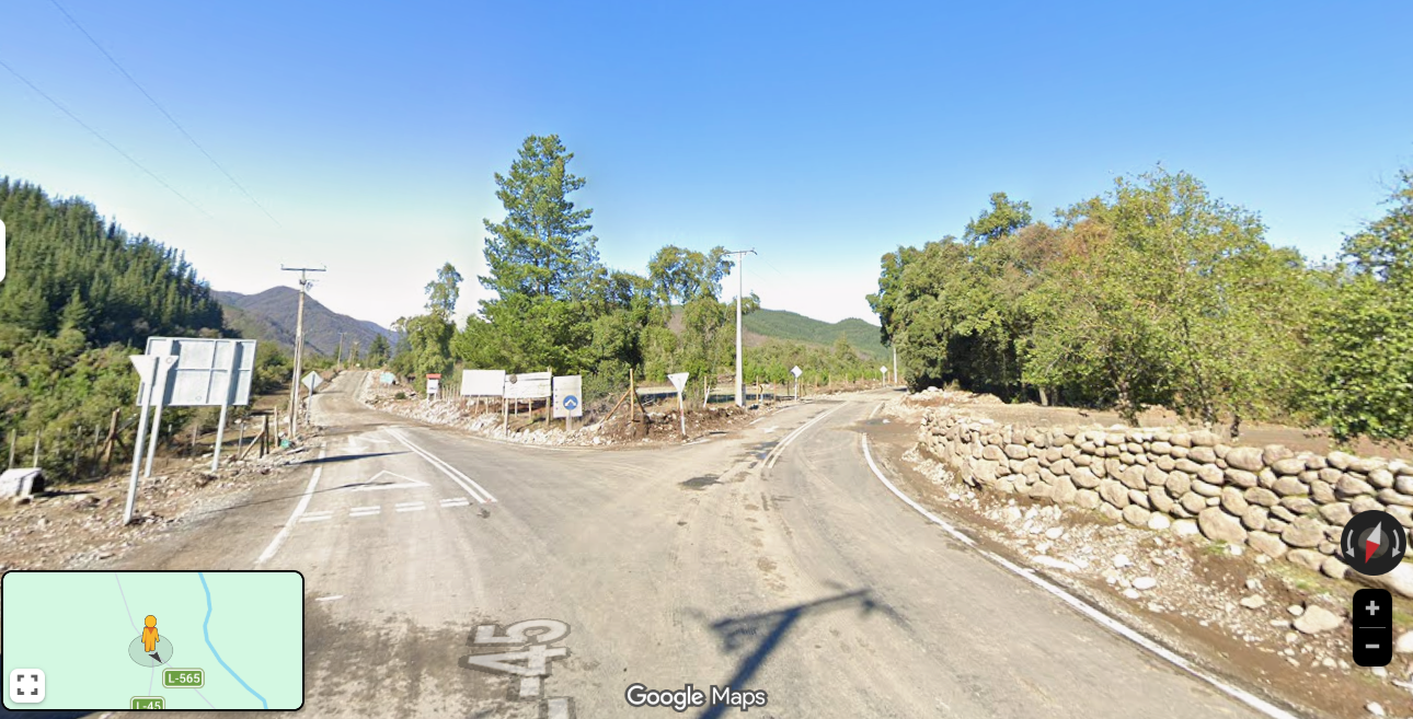 Camino rural con señales de tránsito, árboles y colinas verdes, cielo despejado.