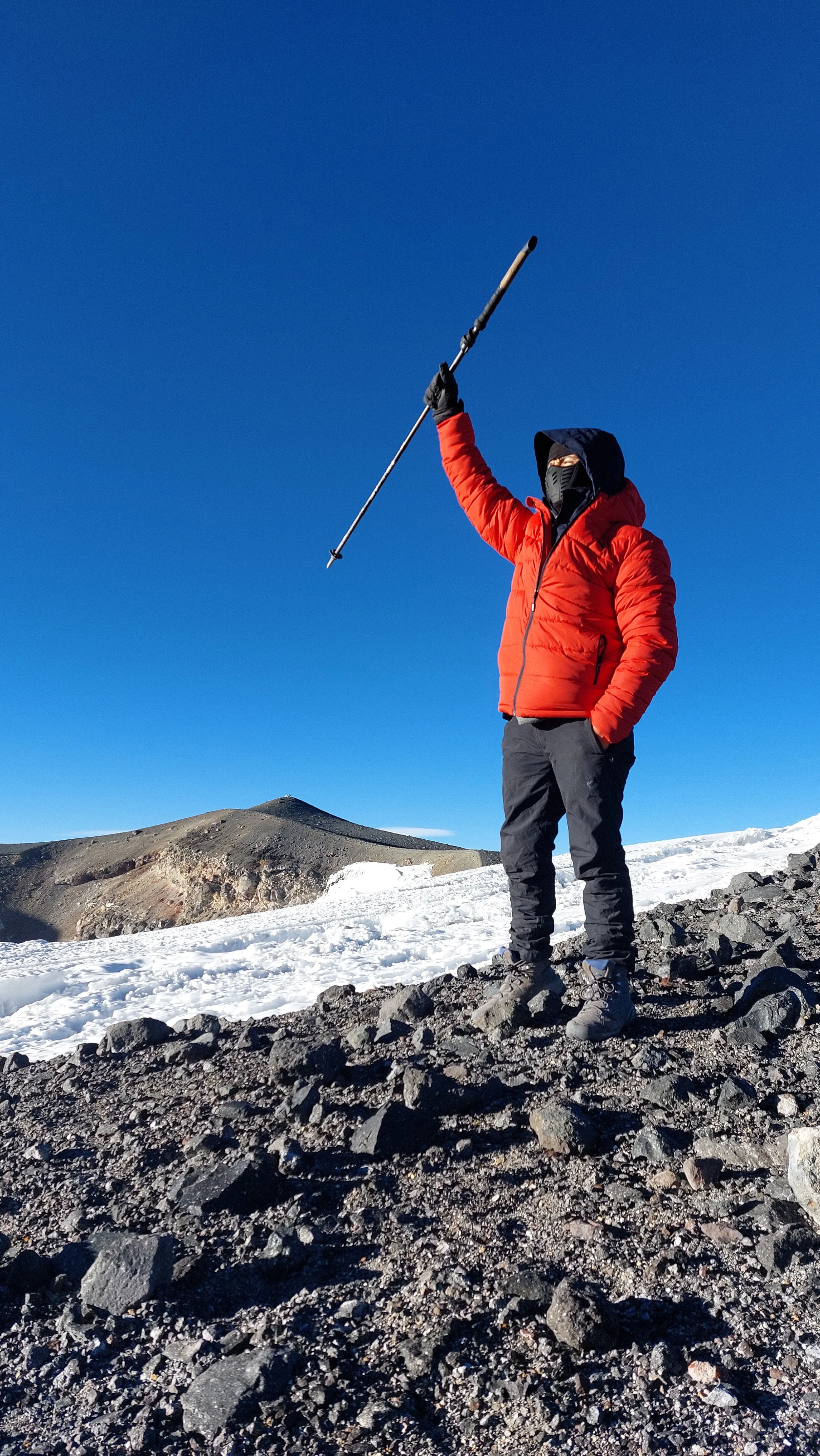 Persona con ropa abrigadora en un paisaje de nieve y volcán en el fondo, levantando un bastón de esquí hacia el cielo azul.