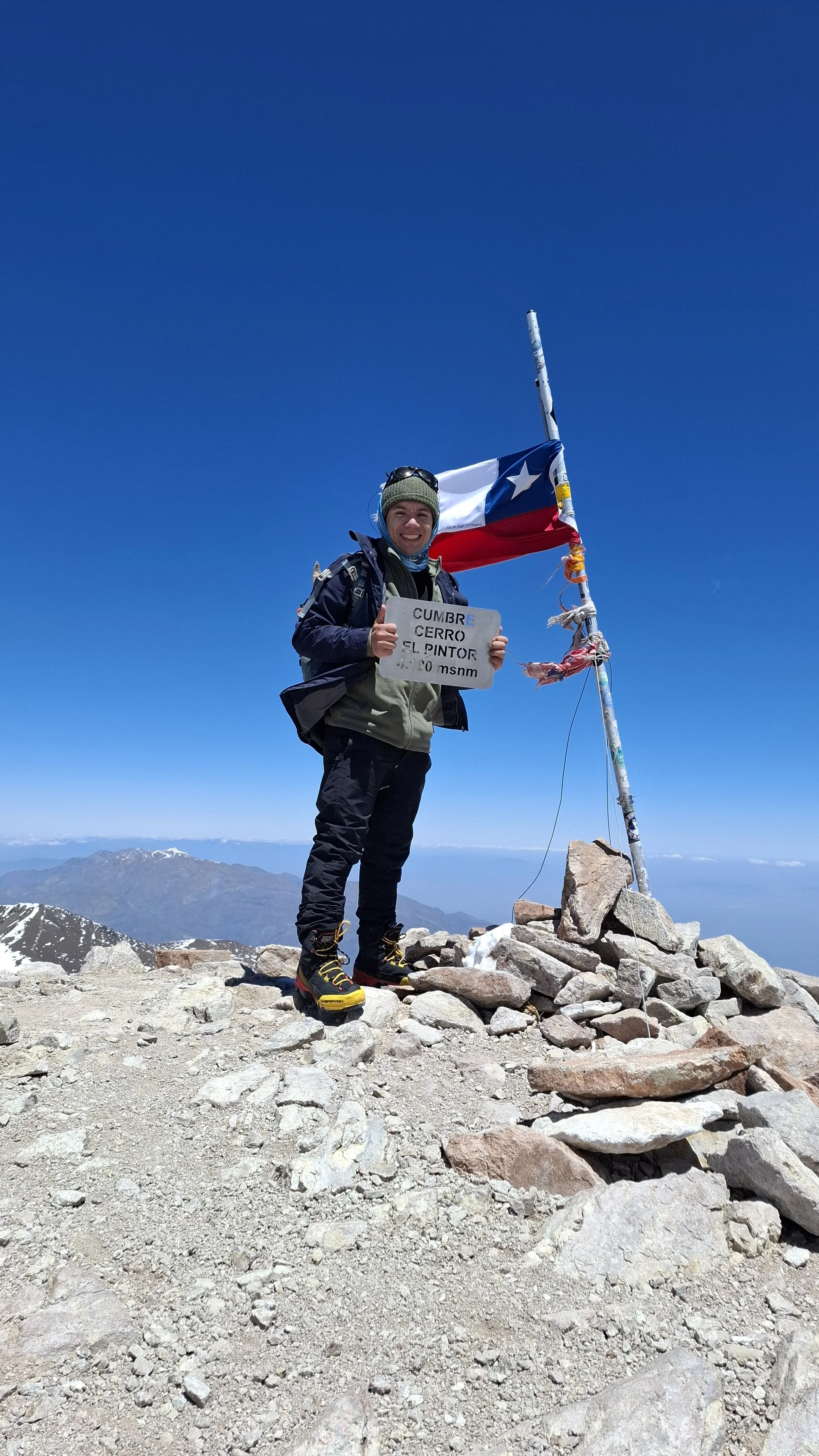 Persona en la cima de una montaña, sosteniendo un cartel que dice "Cumbre Cerro El Pintor 4,180 m snm", con una bandera chilena en una caña y un fondo de picos montañosos y cielo azul.