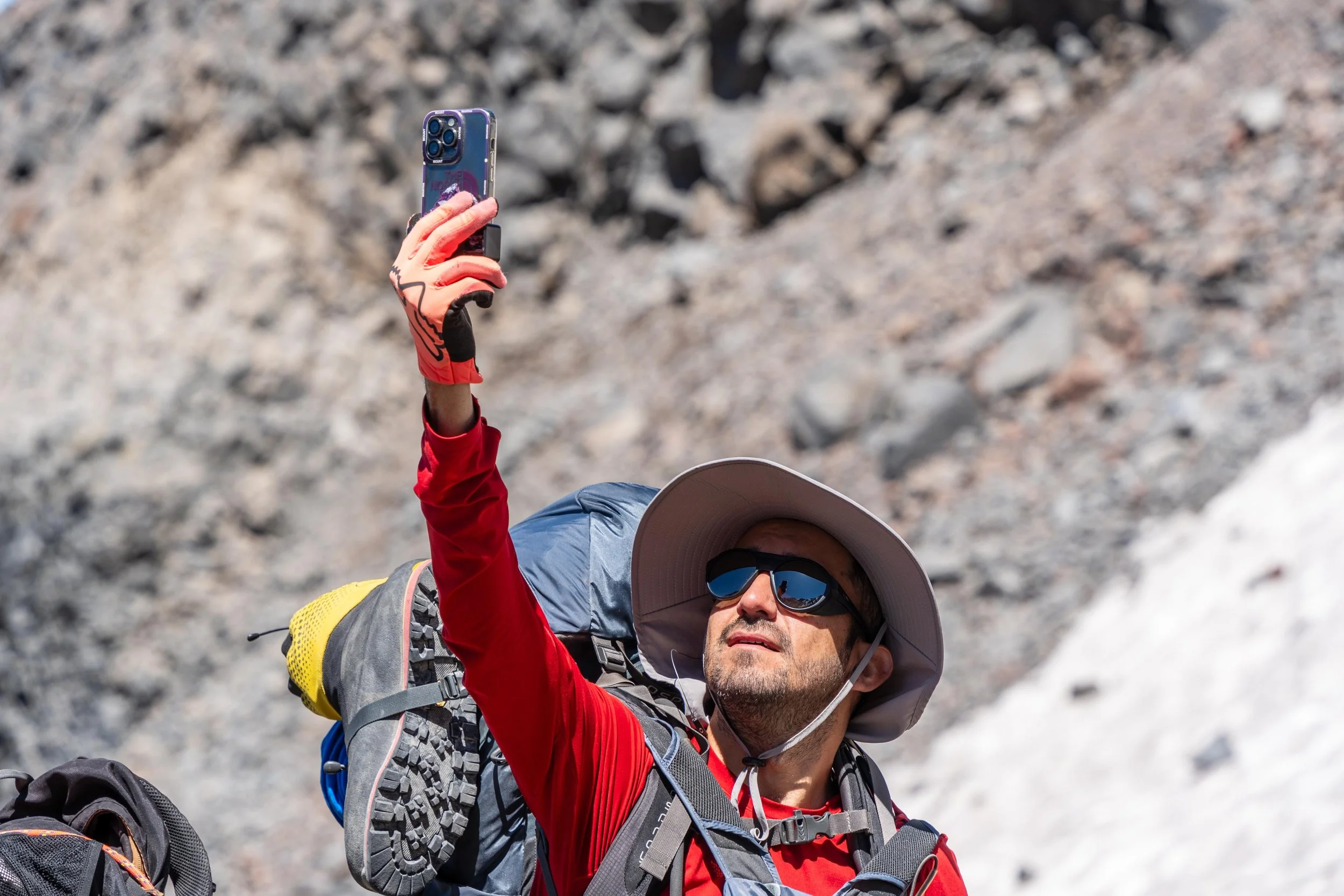Persona con gorra, gafas y guantes rojos tomando una selfie con teléfono móvil en un entorno rocoso.