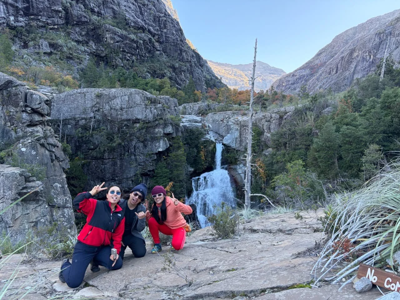 Tres personas posando en un paisaje de montaña con cascada, rocas y árboles, cerca de una señal que dice "No Com."