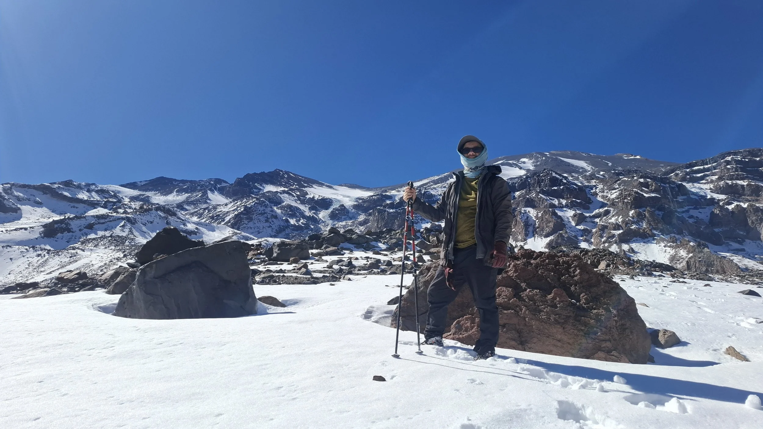 Persona vestido con ropa de abrigo, sombrero, gafas de sol y guantes sosteniendo bastones de esquí en un paisaje de montañas nevadas y cielo azul claro.