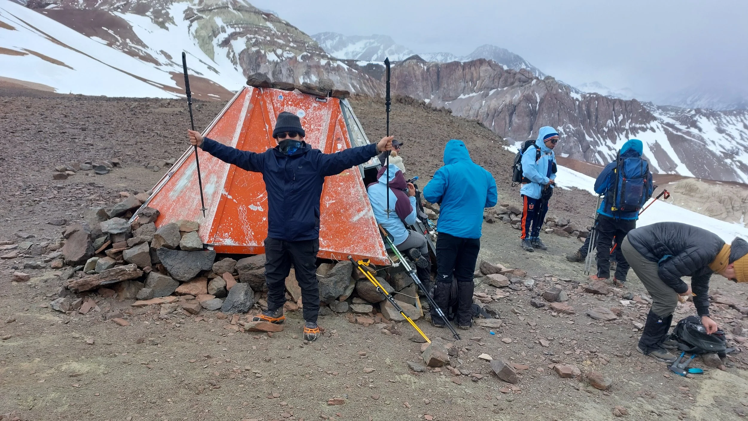 Grupo de personas haciendo senderismo en un paisaje montañoso con nieve y rocas, algunos descansando y otros organizando su equipo en el terreno.