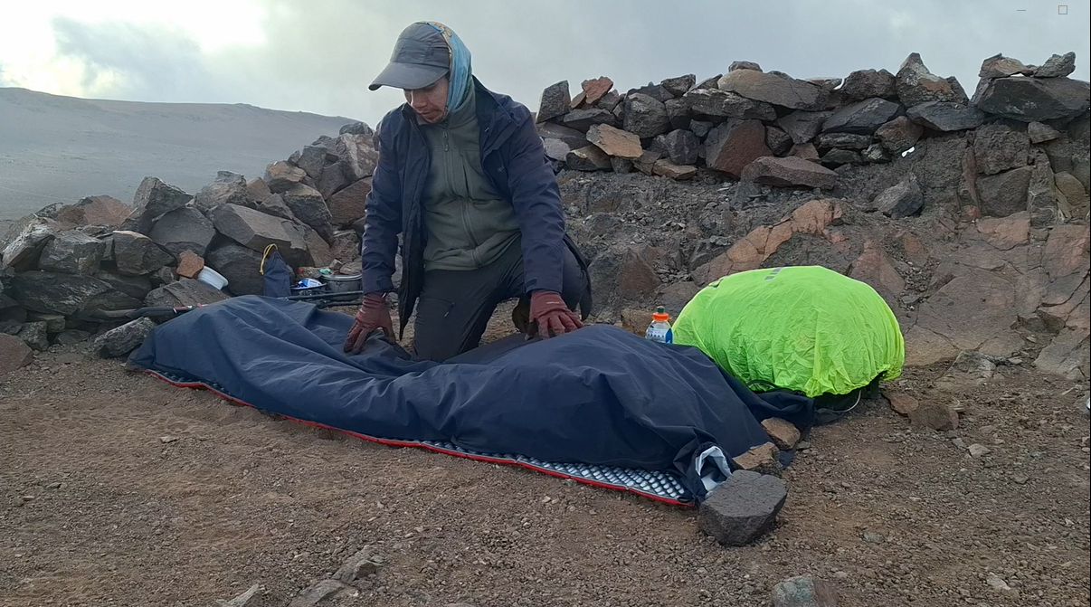 Persona preparando un saco de dormir en un paisaje de montaña rocoso con cielos nublados.