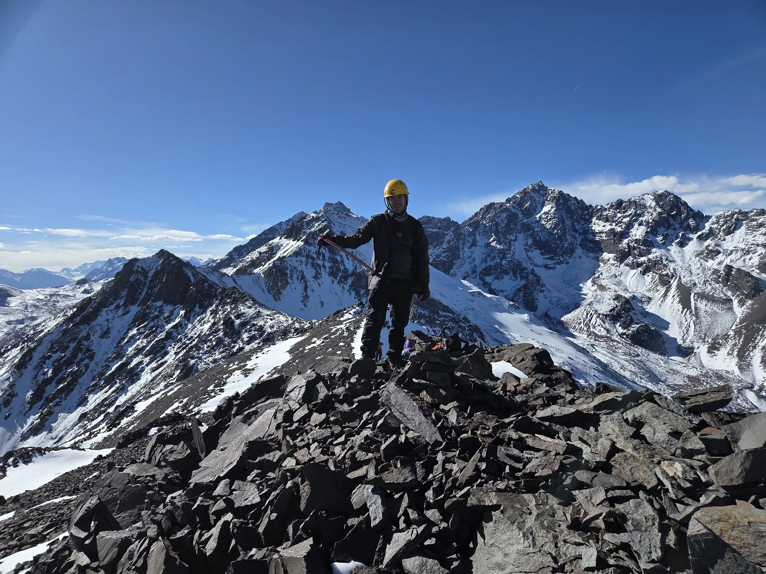 Persona en ropa de montaña con casco y bastón de caminata en una cima nevada, rodeada de picos de montañas y rocas, con un cielo despejado y la luna visible.