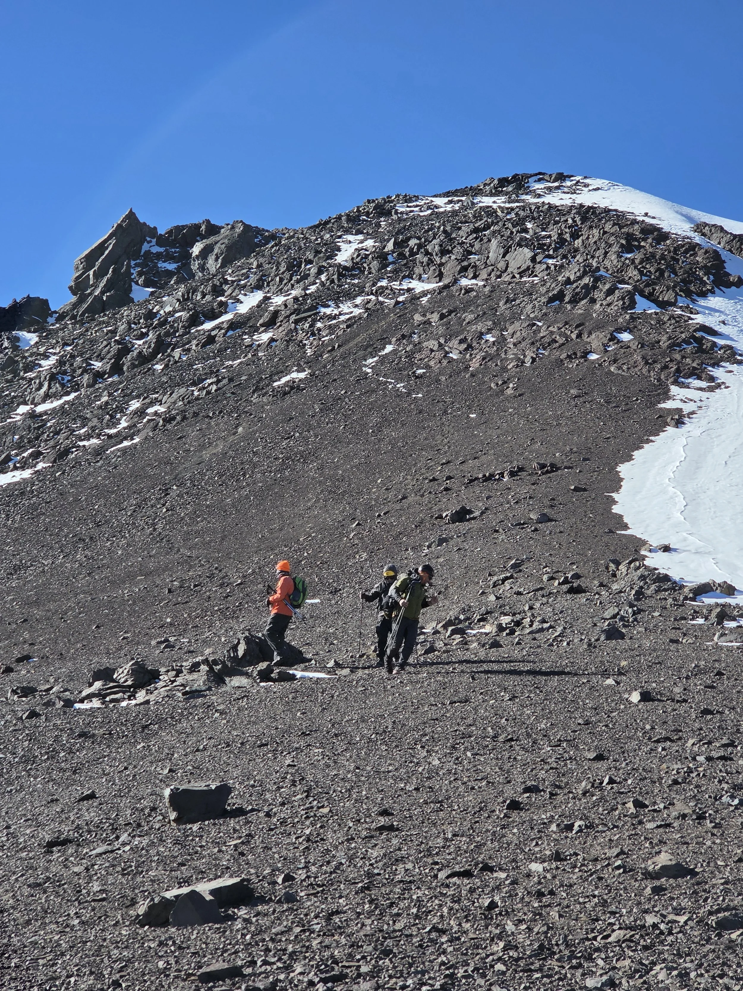 Tres excursionistas en la ladera de una montaña rocosa con nieve en la cima y un cielo despejado.