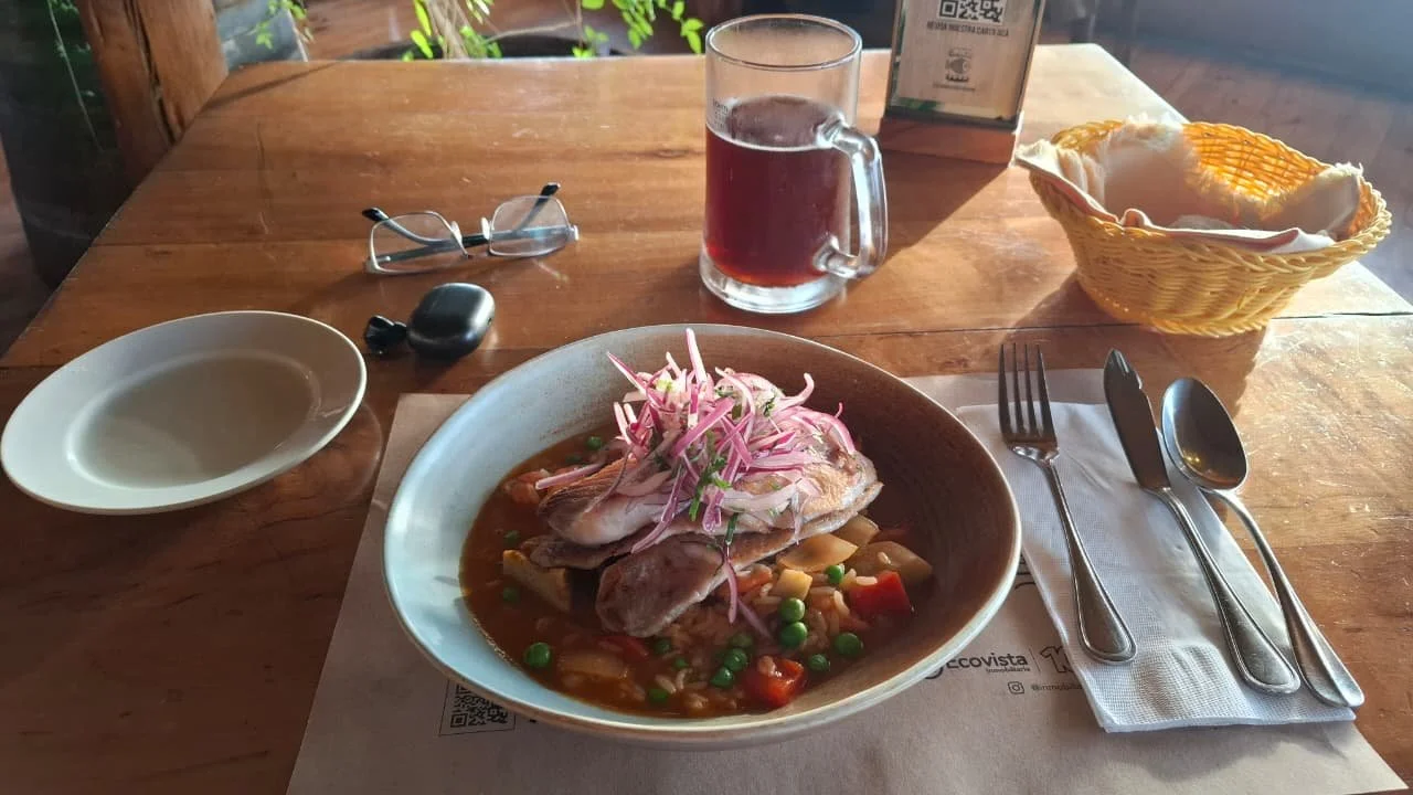 Plato con pollo, arroz, verduras y cebolla morada en un platón, acompañado de un vaso de bebida y un cesta con pan, en una mesa de madera en un restaurante.
