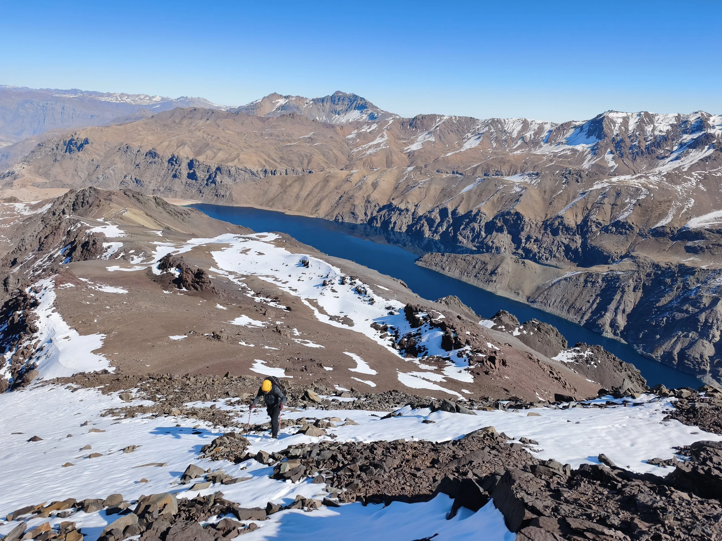 Una persona con casco amarillo y ropa de montaña caminando en un terreno rocoso y nevado en una cordillera montañosa con lagos y picos cubiertos de nieve en el fondo.
