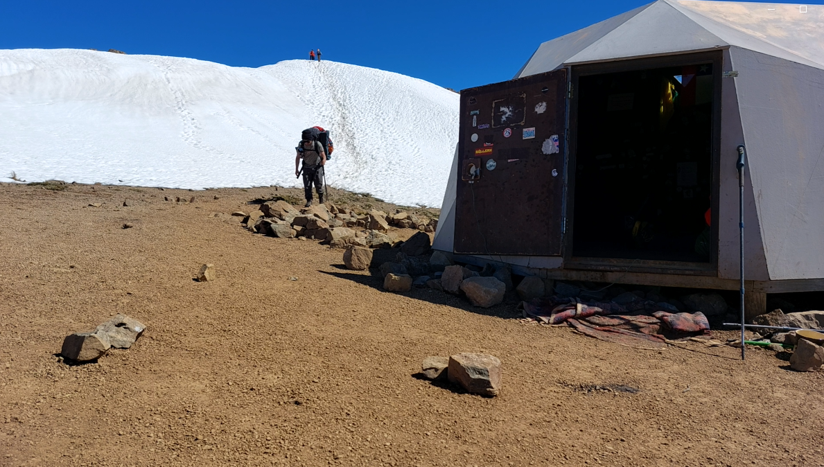 Personas haciendo senderismo en una montaña con nieve, cerca de una estructura de camping o refugio de lona en un paisaje árido.