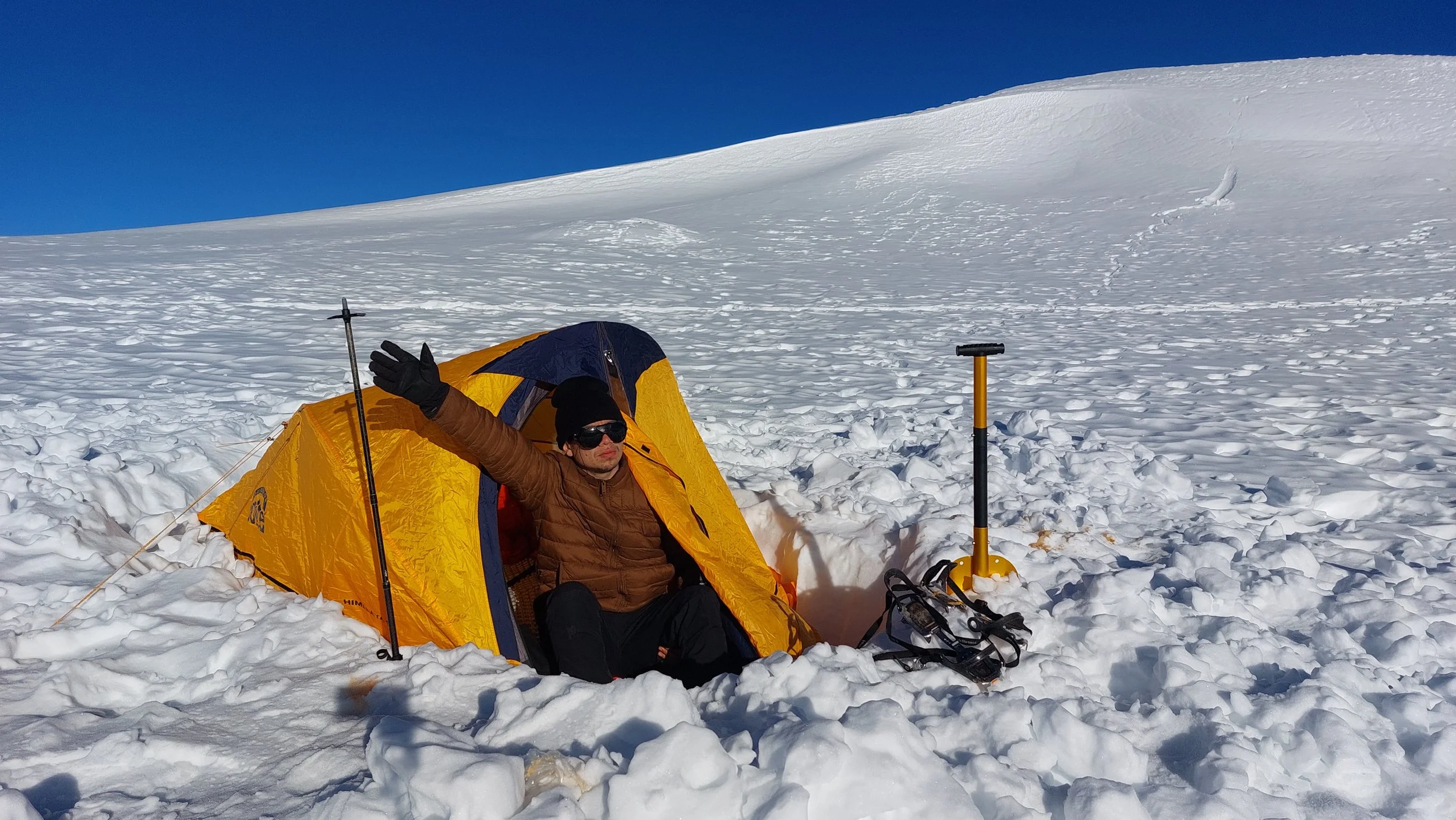 Persona en ropa de abrigo en una tienda de campaña amarilla en la nieve, con un pico y una pala en el hielo, en un paisaje de nieve y cielo despejado.