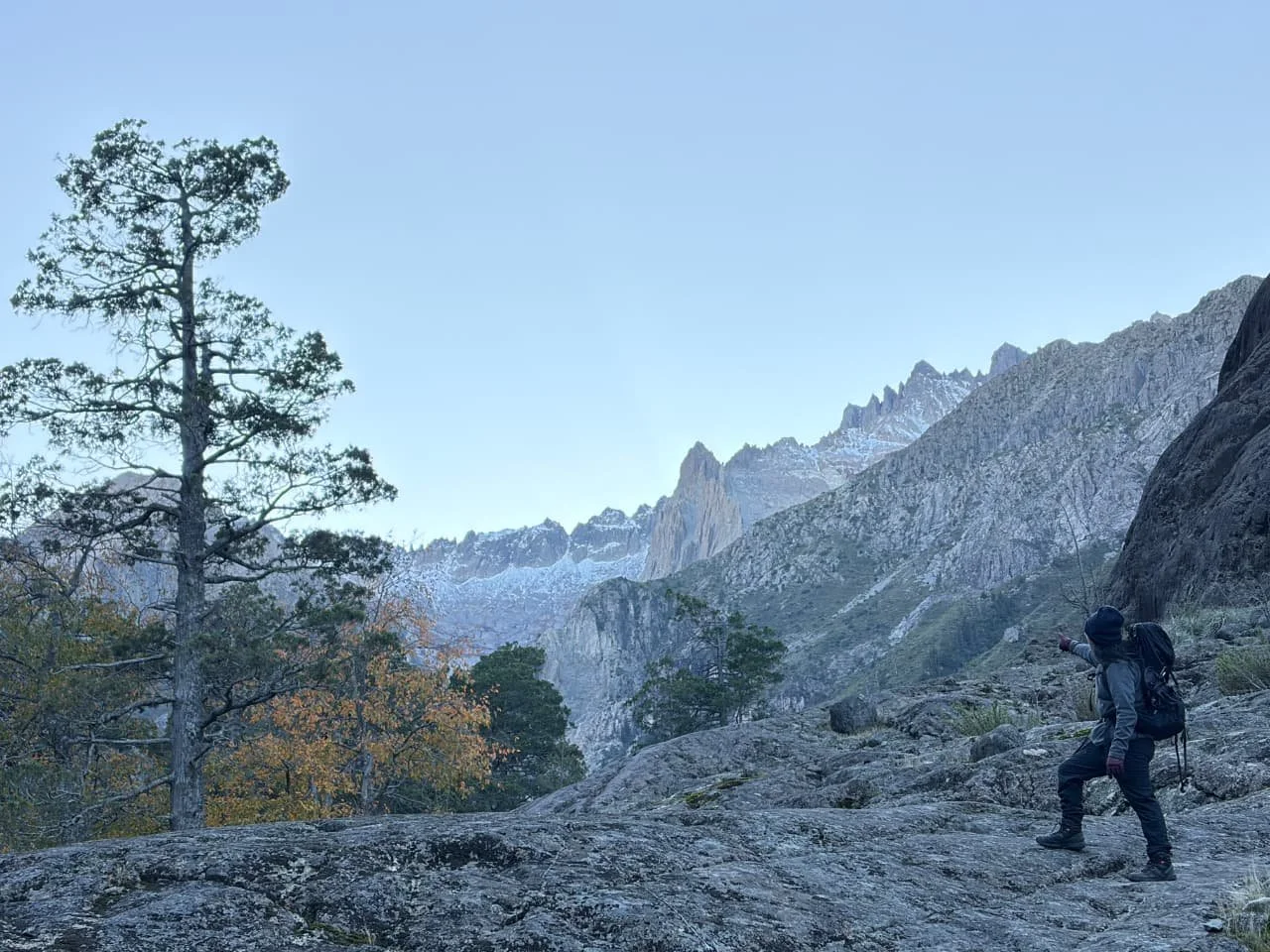 Persona haciendo senderismo en un paisaje montañoso y rocoso con árboles y picos nevados en el fondo.