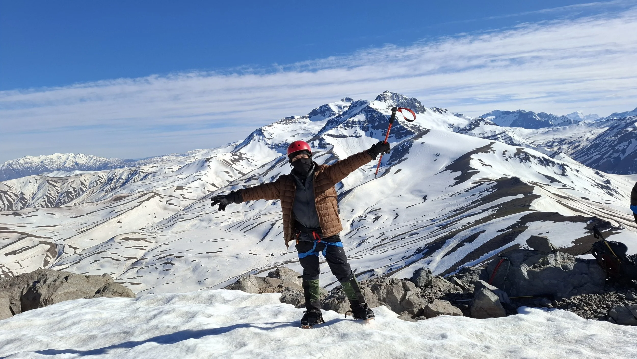 Persona con equipo de montañismo en la cima de una cordillera nevada, levantando los brazos en señal de triunfo con un paisaje de montañas y cielo azul de fondo.