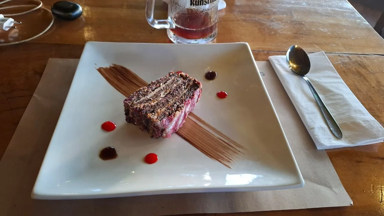 Postre de pastel de varios capas decorado con salsa de chocolate y pequeñas gotas de sirope rojo, en un plato blanco rectangular con decoración de salsa de chocolate y un trago detrás.
