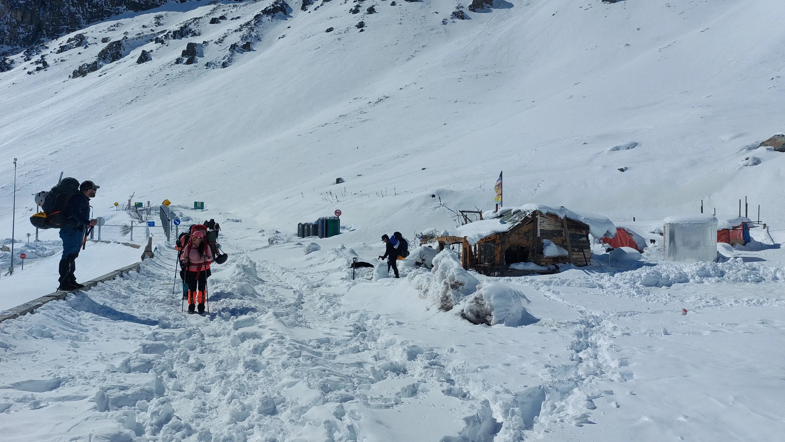 Personas en un entorno de montaña nevada, con refugios y señalización, preparándose para actividades al aire libre en la nieve.