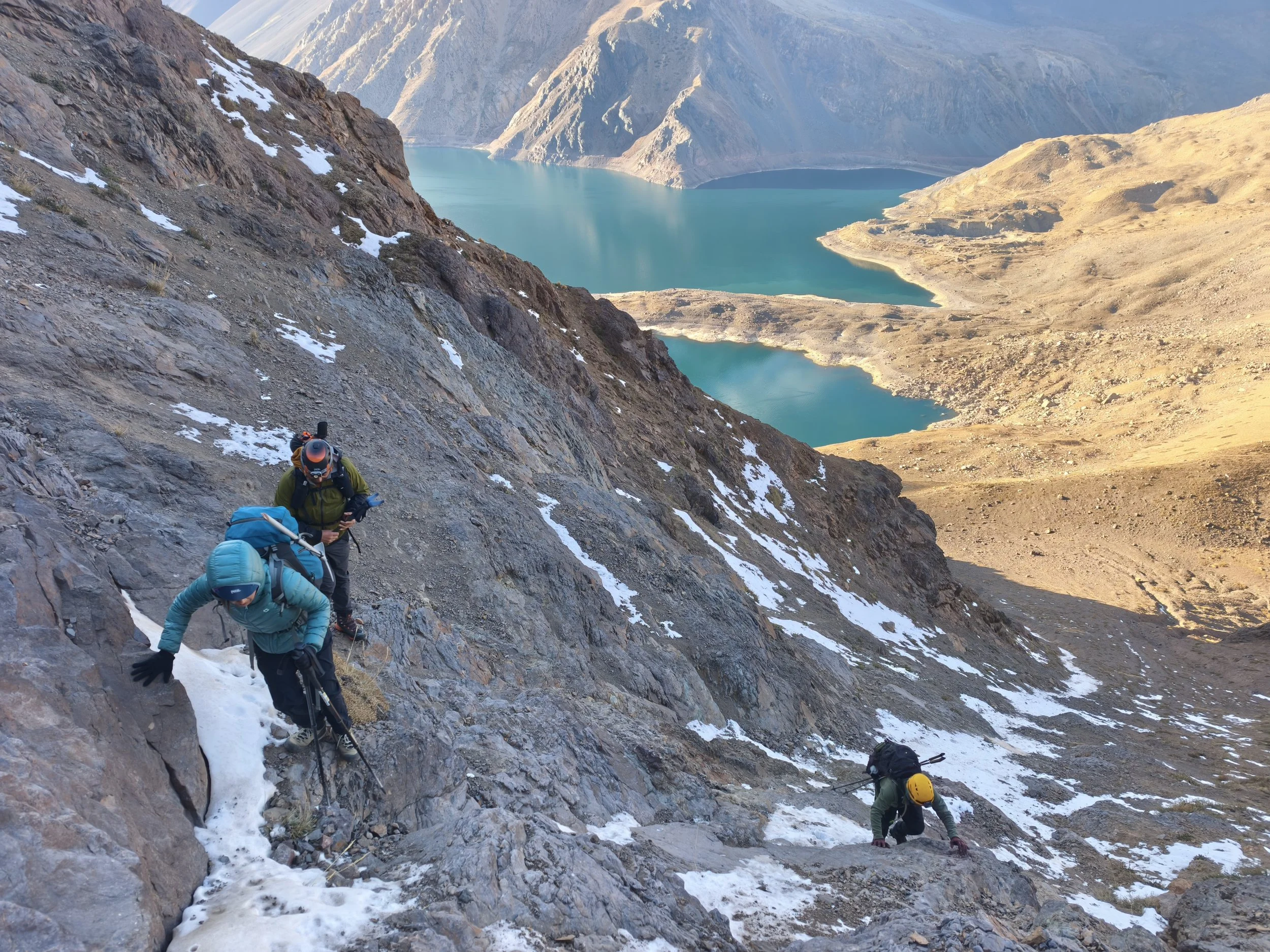 Montañistas ascendiendo una pendiente empinada de roca y nieve, con un lago de color turquesa y montañas en el fondo.