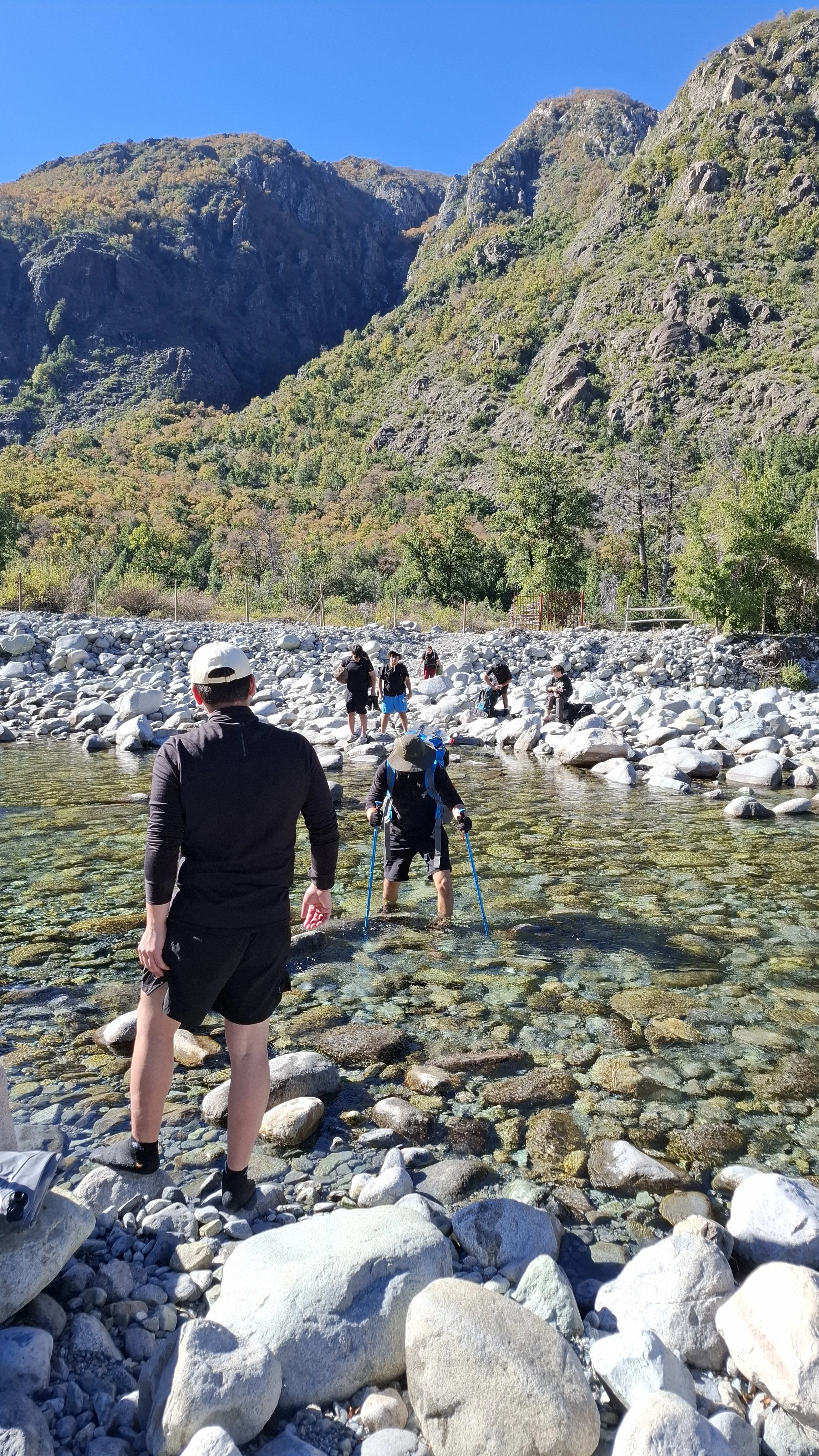 Grupo de personas cruzando un río de aguas claras y piedras, rodeados de montañas verdes y rocosas con un cielo azul despejado.