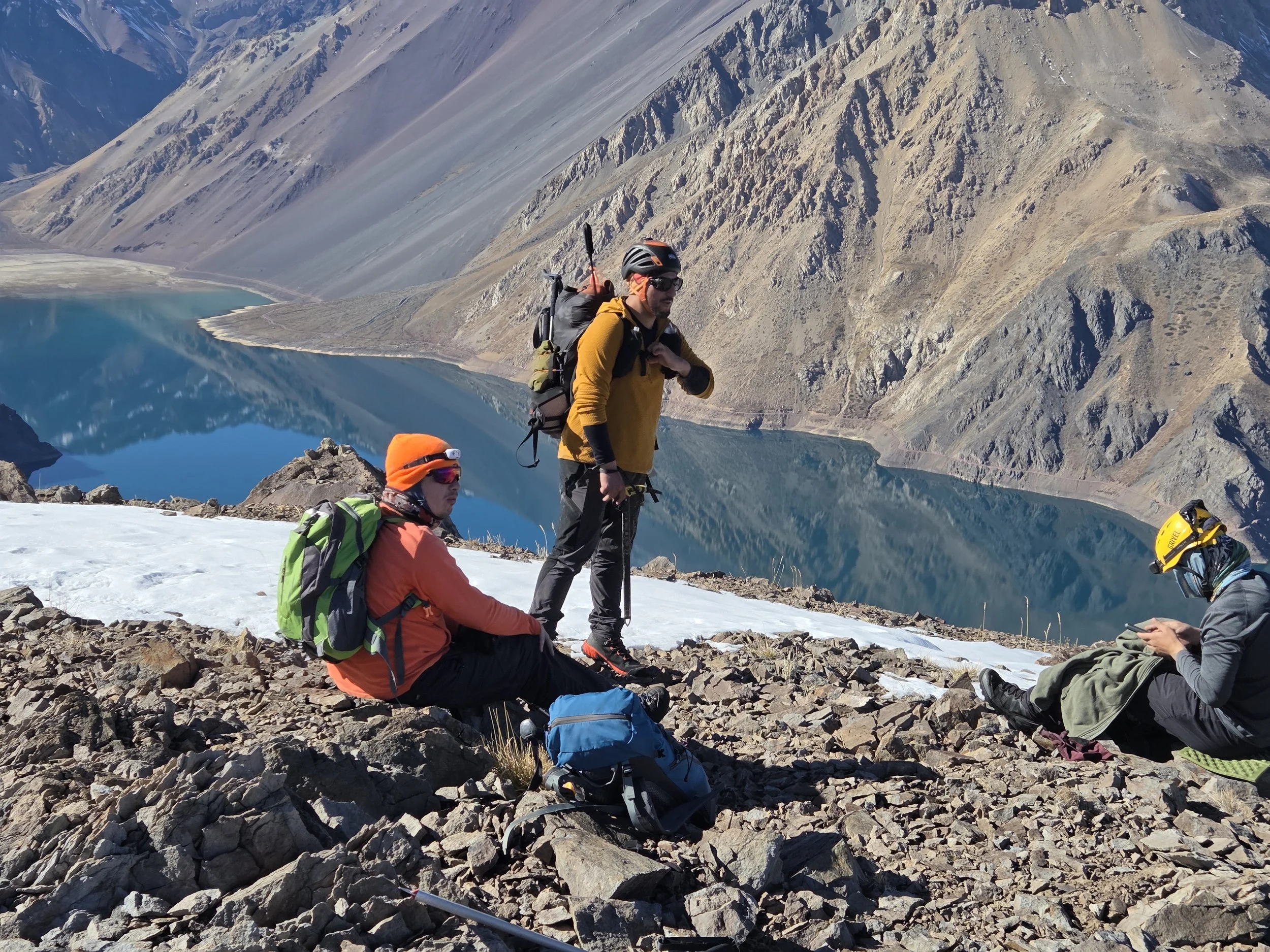 Grupo de senderistas descansando en una zona rocosa junto a un lago de aguas azules y montañas de tonos grises y marrones en un paisaje de montaña.