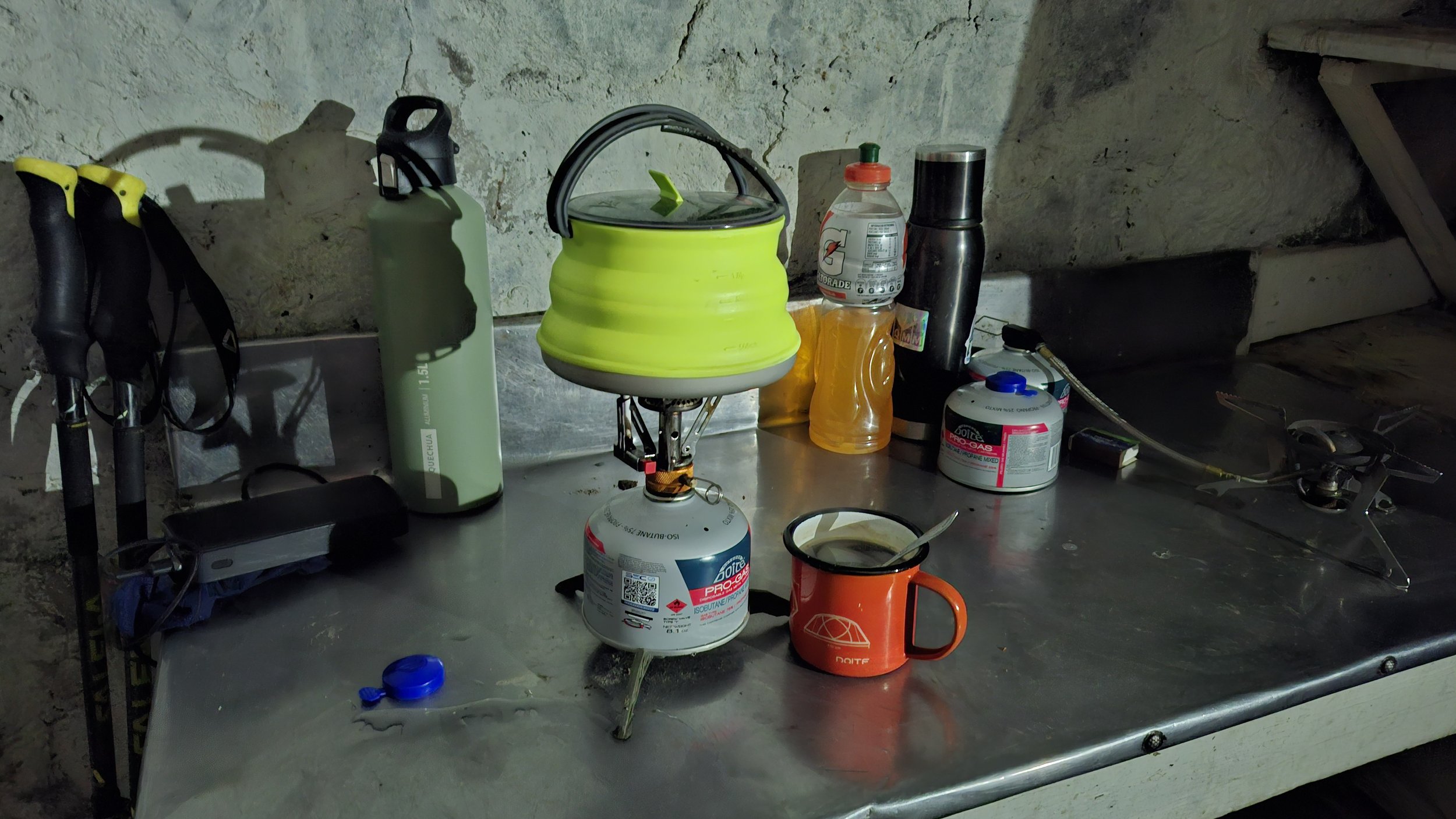 Mesa de trabajo en una pared de piedra con varios utensilios y equipo de camping, incluyendo una lámpara portátil, botellas de agua, una cafetera, una taza naranja, y otros objetos diversos.