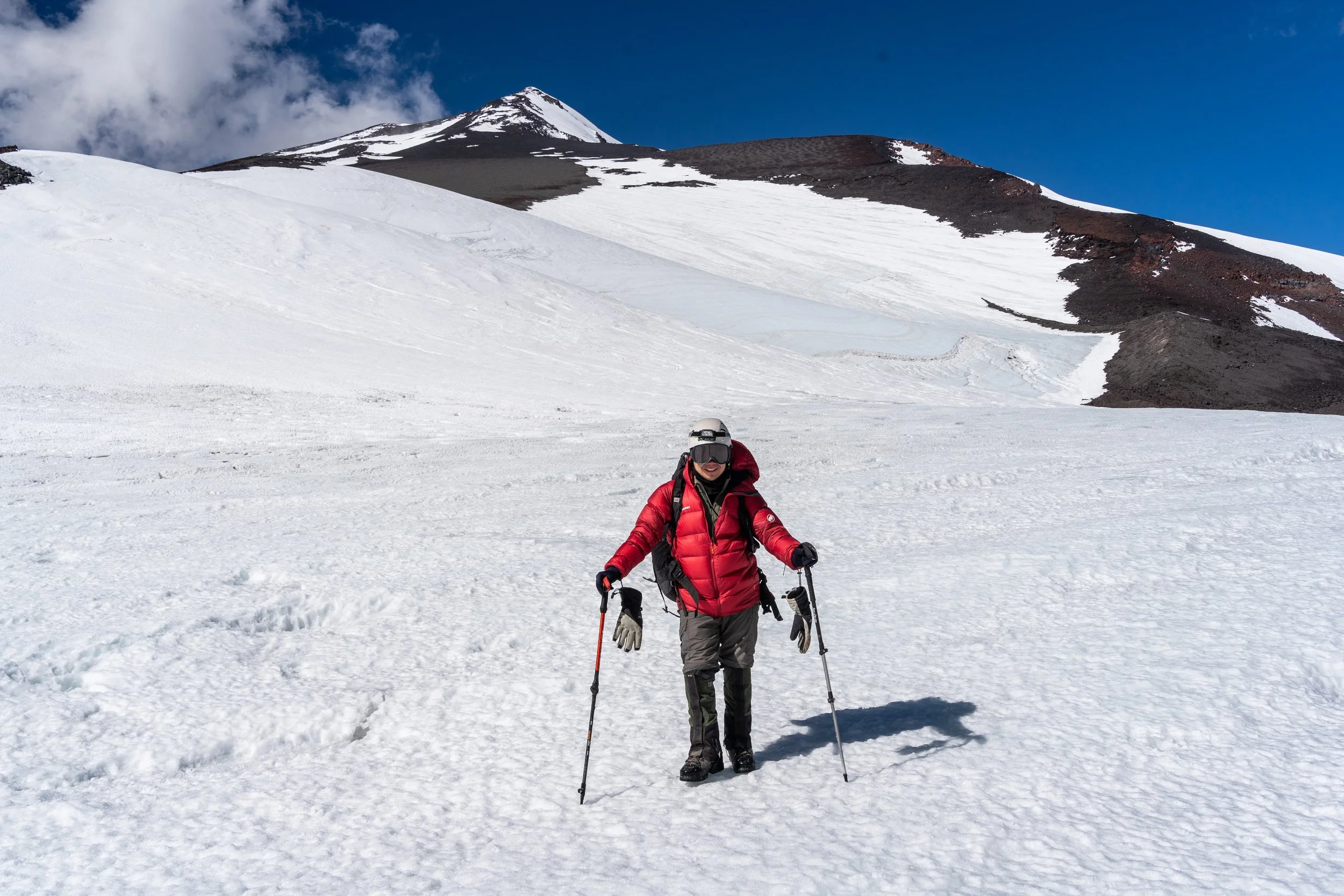 Personas caminando en nieve en una montaña con cielo azul y nubes, con vestimenta de abrigo y equipo de esquí.