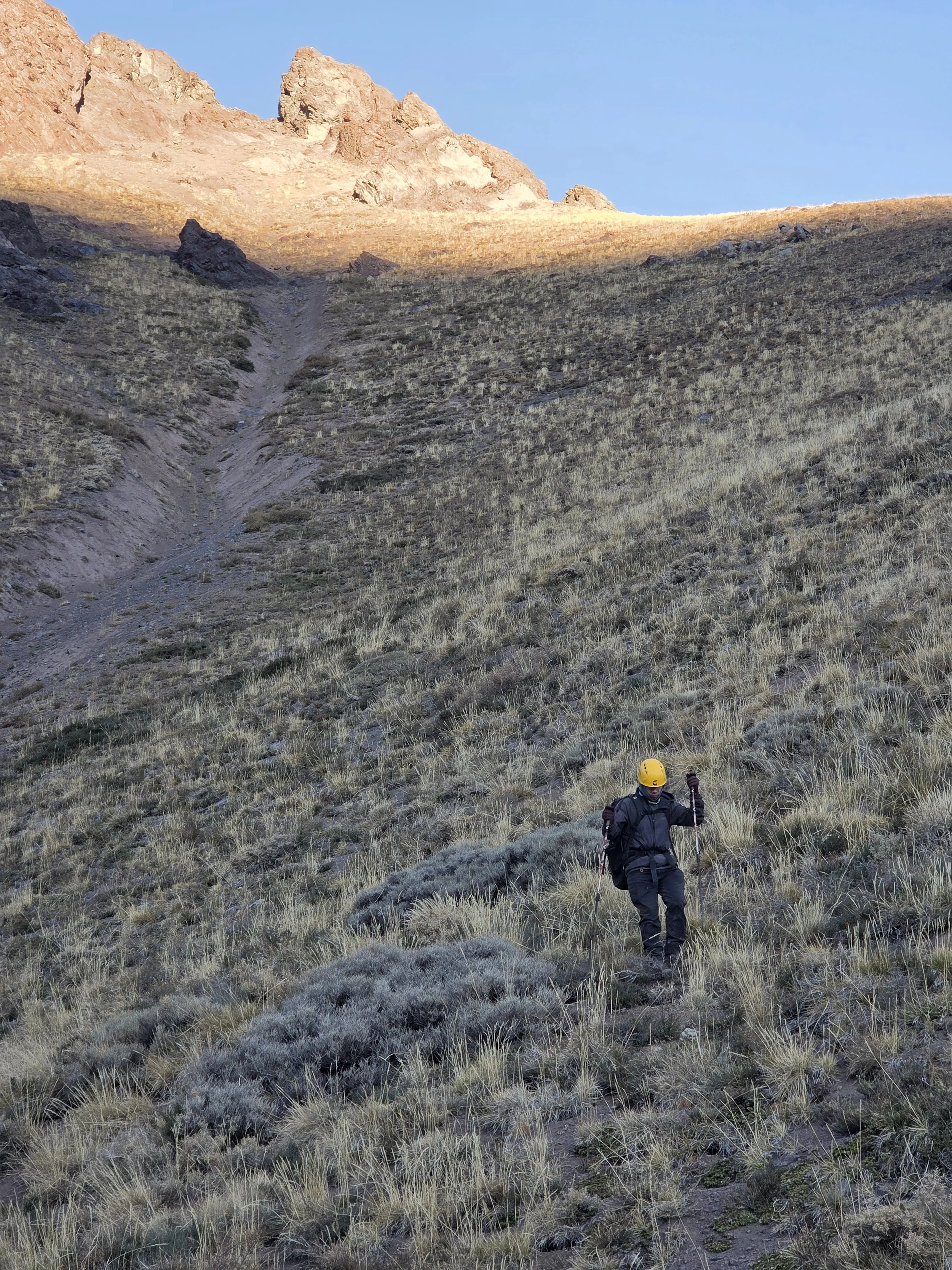 Persona caminando por un sendero en una ladera montañosa con rocas y césped seco, con una montaña en el fondo y un cielo despejado.