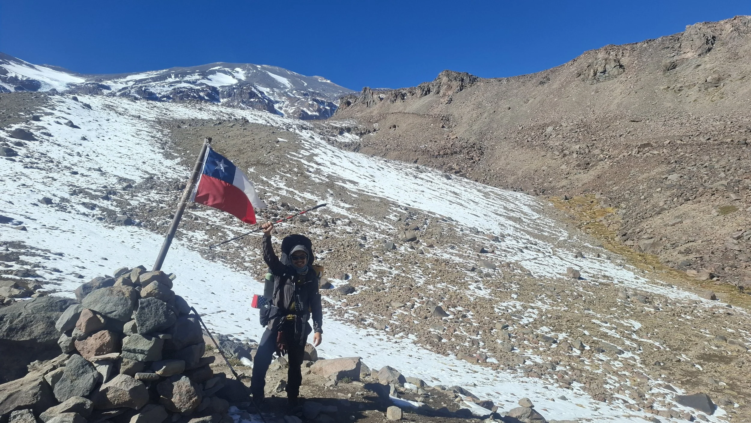 Persona sonriendo en la cima de una montaña, sostiene una bandera de Chile, rodeada de roca y nieve con un fondo de montañas y cielo azul.