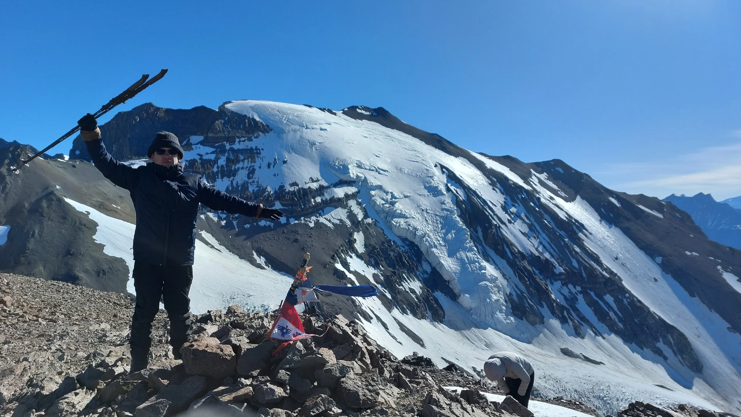 Persona en la cima de una montaña nevada con un pico y banderas, vestida con ropa de abrigo, en un entorno de montaña con hielo y rocas.
