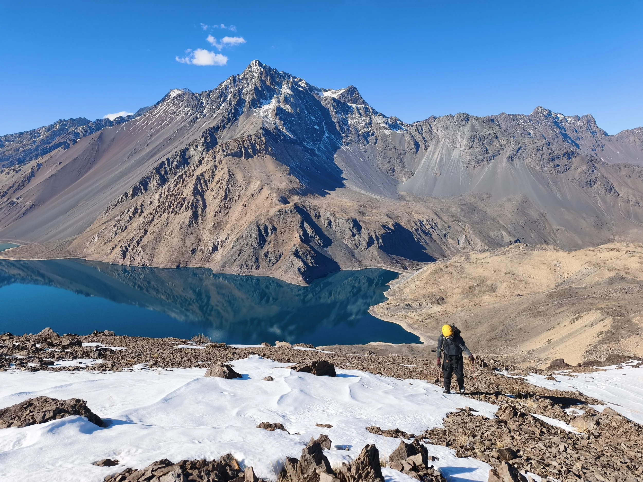 Montaña de gran altura con picos rocosos y nieve en la cima, reflejada en un lago de agua azul. En primer plano, senderista con casco amarillo caminando sobre tierra y nieve.