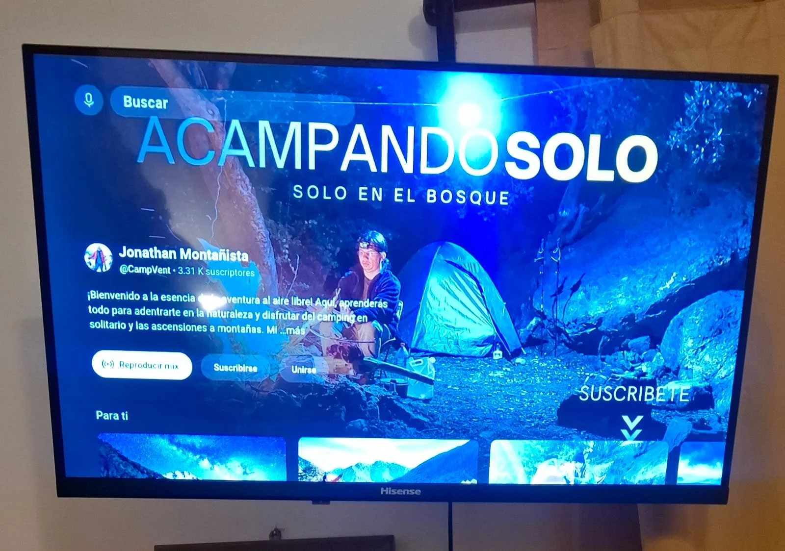 Pantalla de televisor que muestra un canal o perfil de aventura en la naturaleza, con un título grande que dice 'Acampando solo' y un fondo de una tienda de campaña en un entorno de bosque con rocas y árboles.