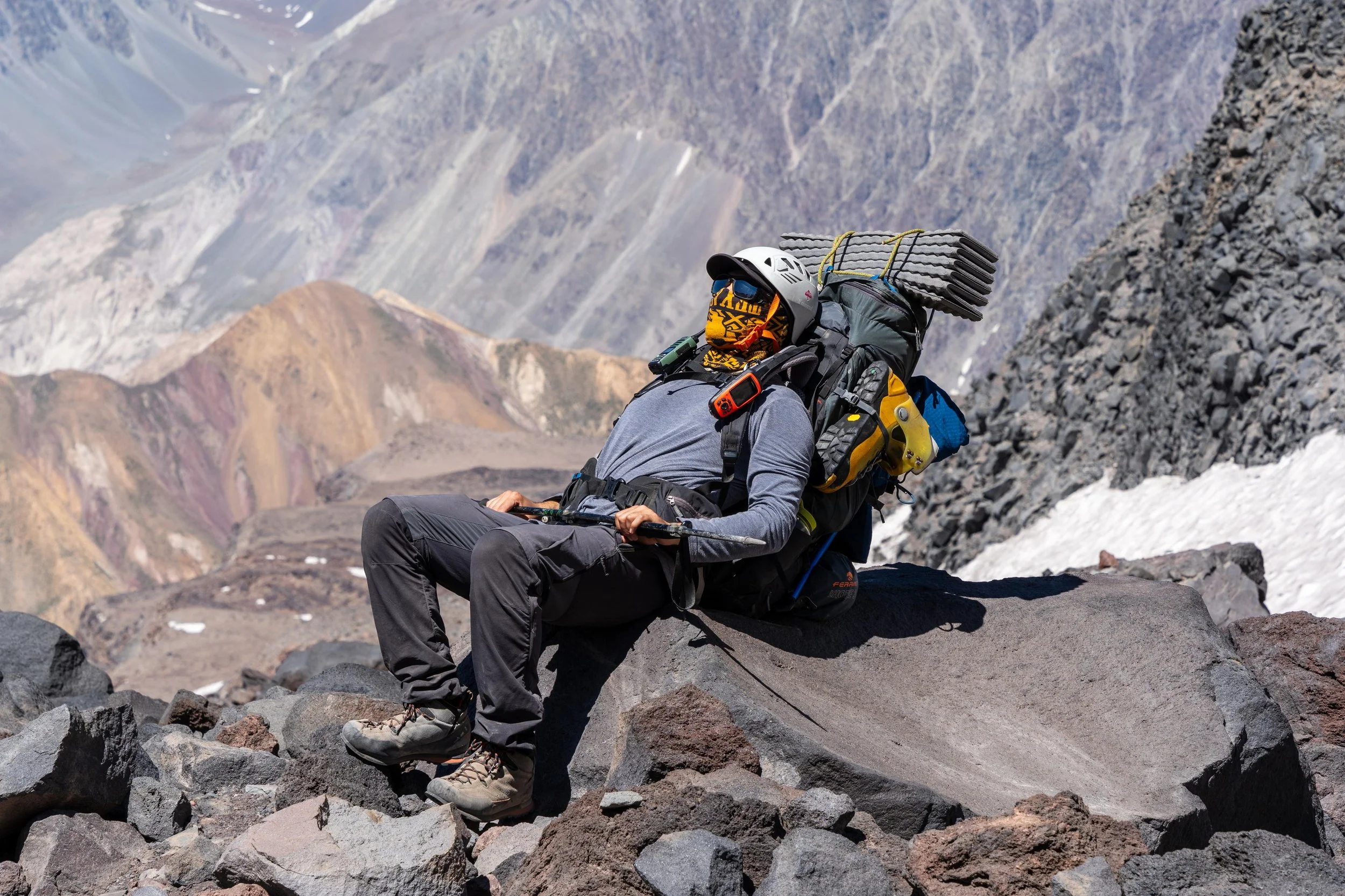 Un excursionista descansando sobre rocas en una zona montañosa con picos nevados en el fondo.