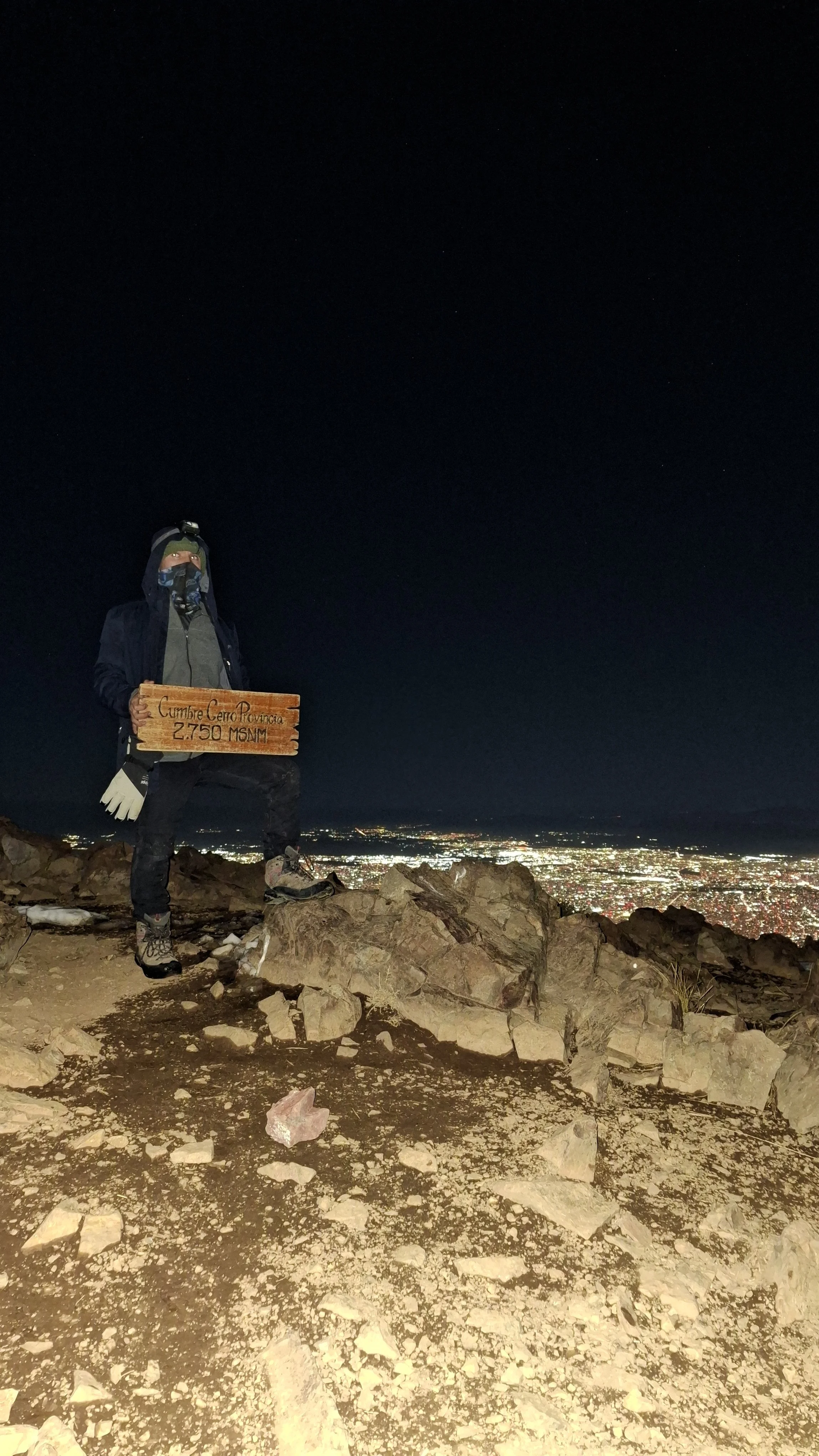 Persona en la cima de una montaña durante la noche, sosteniendo un letrero que dice "Cumbre Cerro Provincia 2,750 MSN" con una vista de la ciudad iluminada en el fondo.