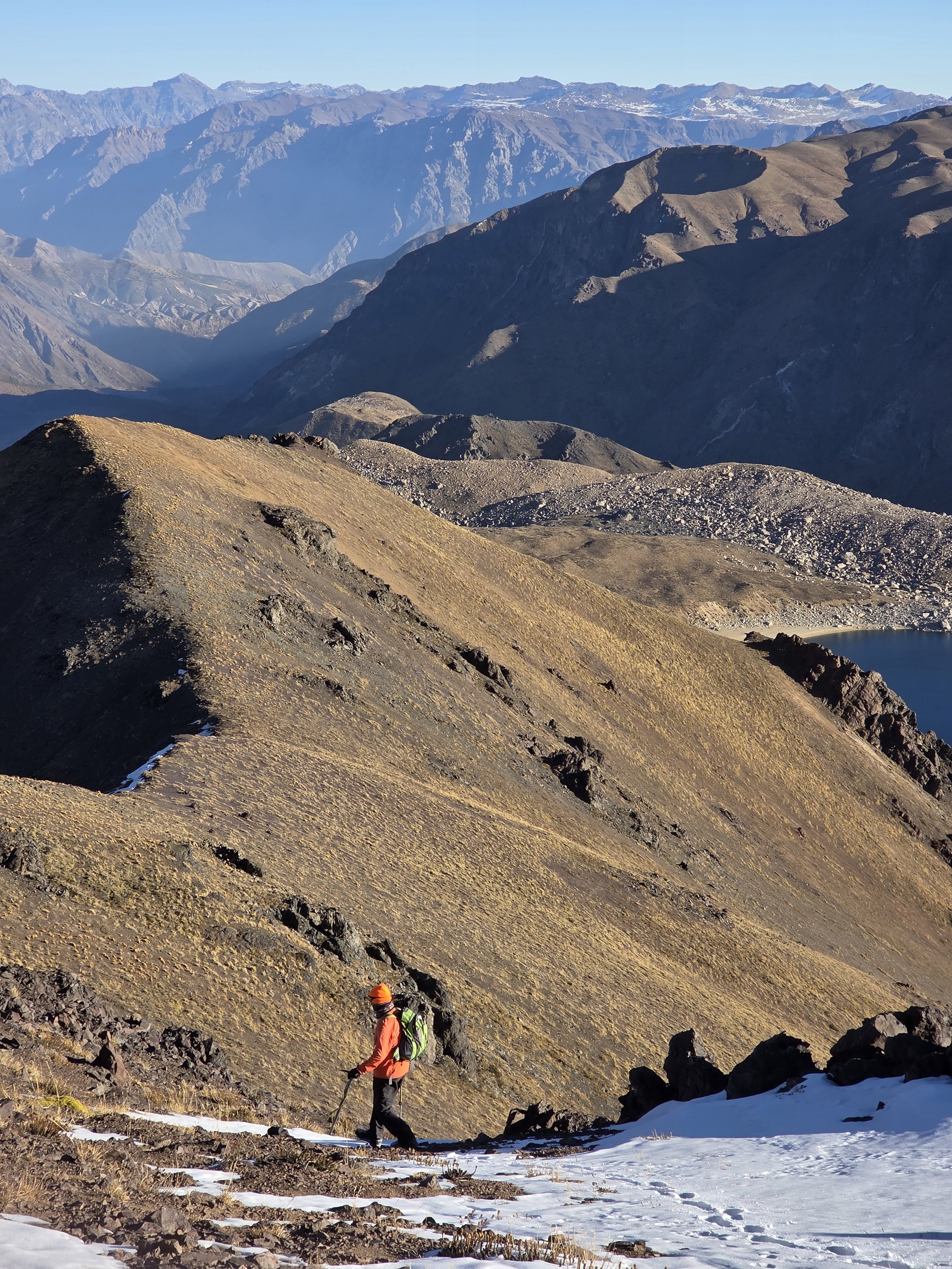 Dos excursionistas en un paisaje montañoso con picos nevados y un lago en el fondo, uno lleva ropa naranja y un gorro. La escena muestra un terreno rocoso y parcialmente cubierto de nieve.