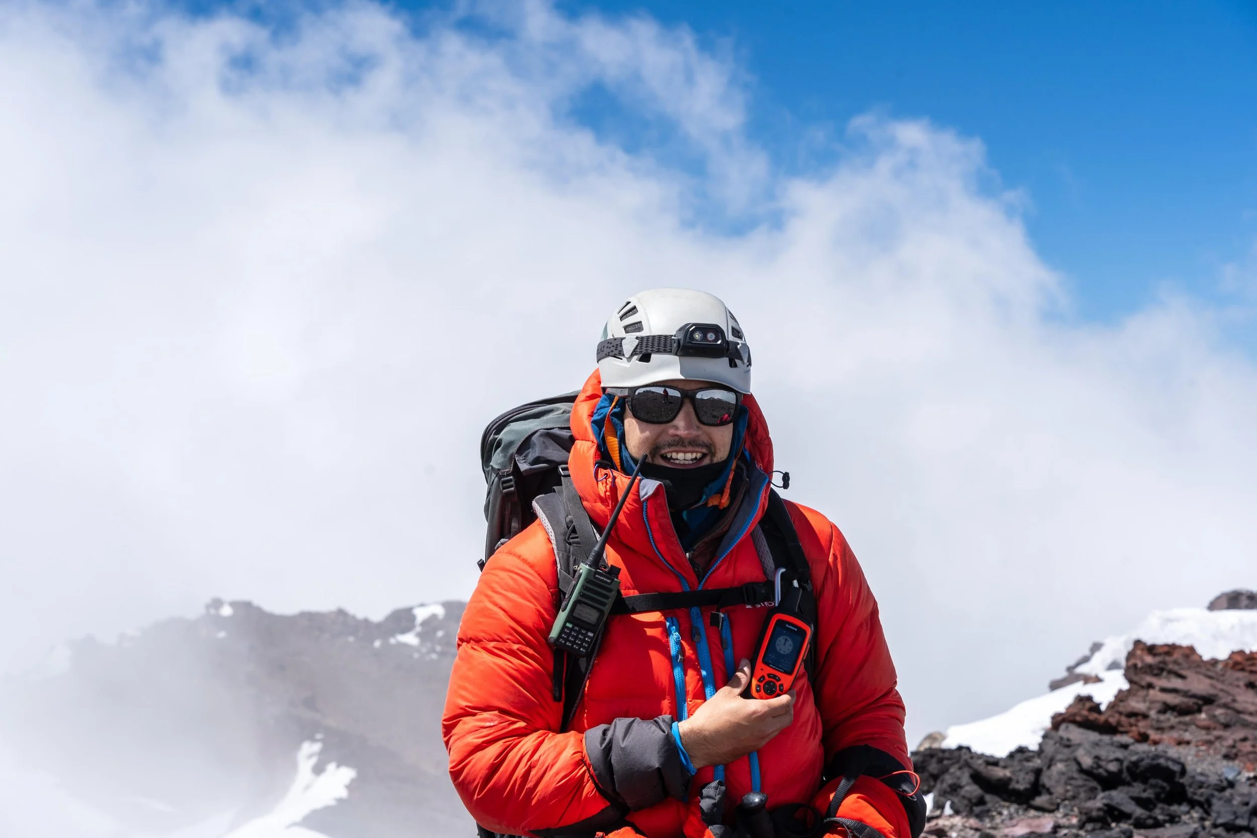Aventurero con equipo de escalada en una montaña nevada, sosteniendo un dispositivo de navegación, con casco y gafas de sol, frente a un cielo azul y nubes.