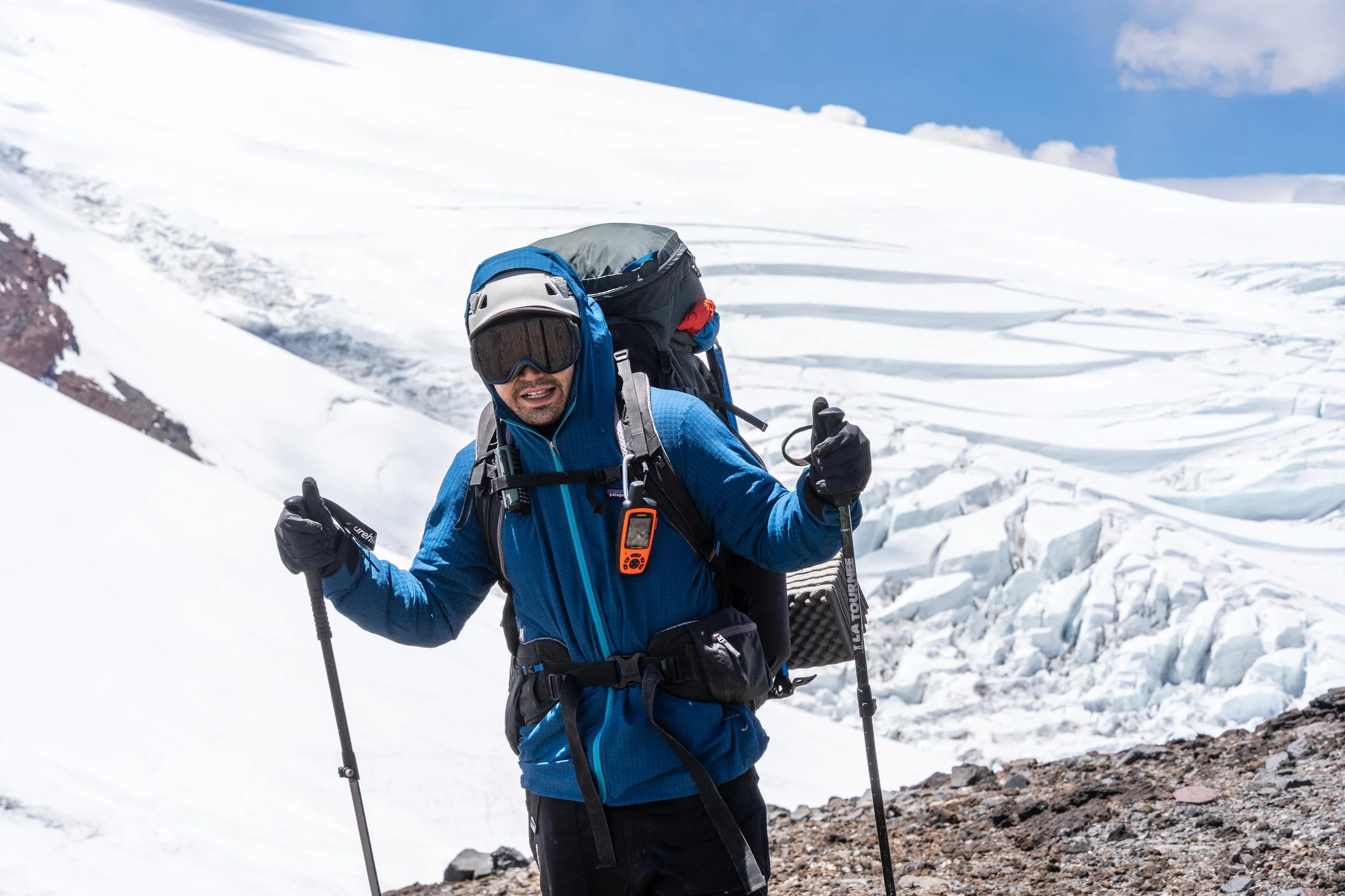 Persona en ropa de abrigo y gafas de protección, sosteniendo bastones de esquí, en un paisaje de nieve y montañas con cielo azul.