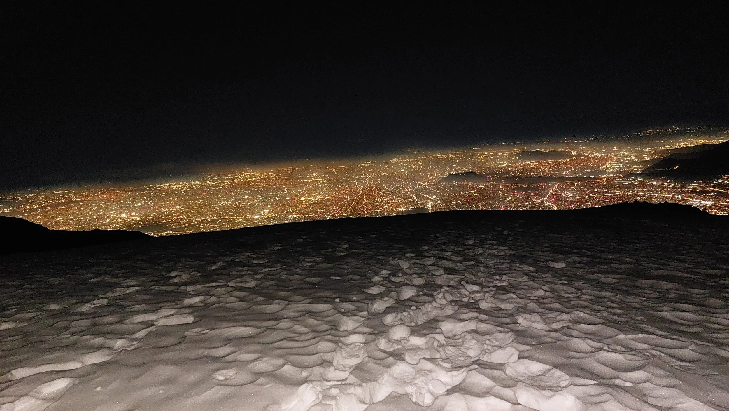 Vista nocturna de la ciudad desde una cima, con un suelo de piedras, iluminada por las luces urbanas y el cielo oscuro.