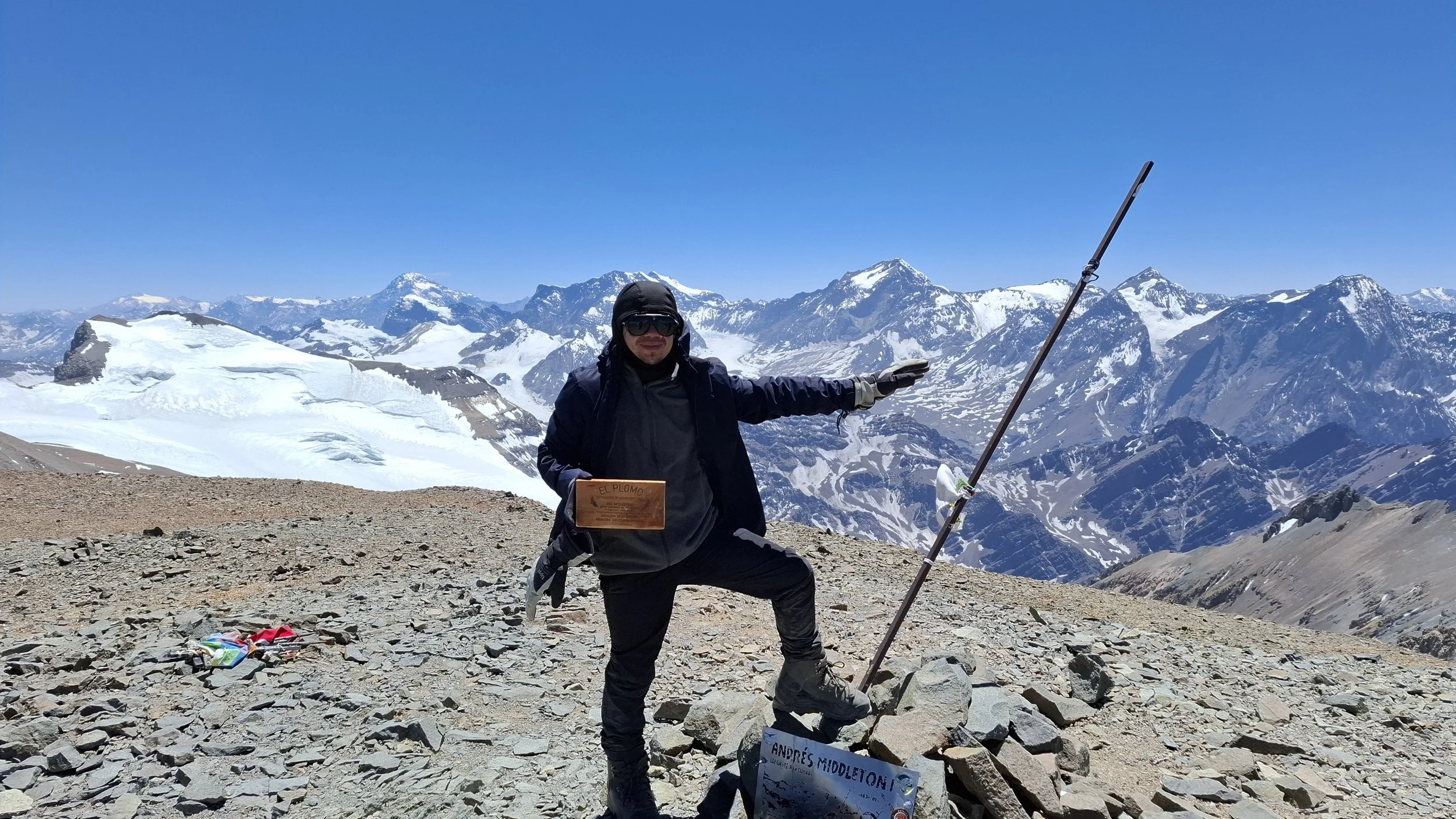 Persona en la cima de una montaña nevada, sosteniendo un palo de senderismo y una placa con su nombre y logotipo, rodeada de picos montañosos y un cielo despejado.