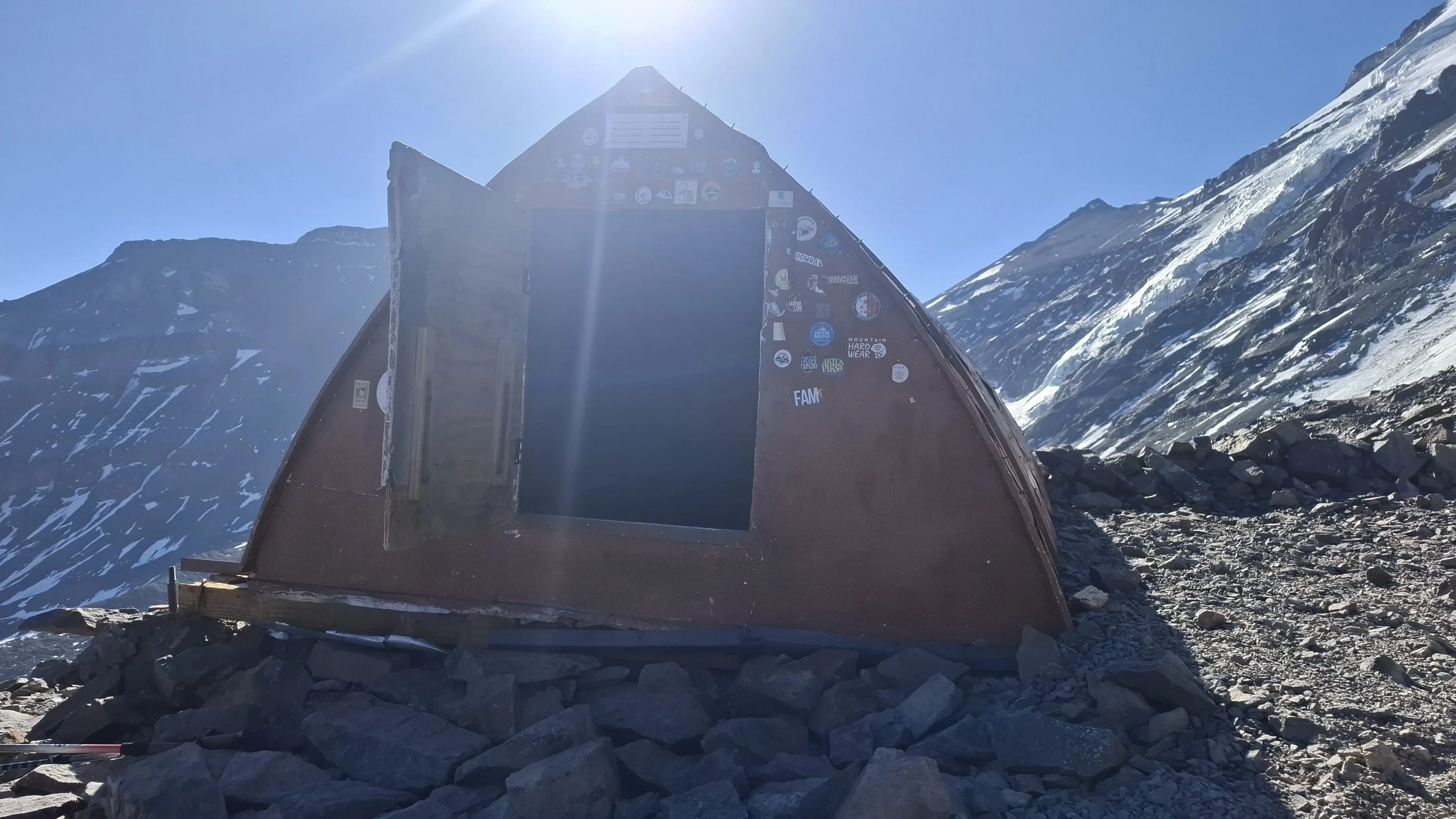 Refugio de montaña en un paisaje nevado con montañas y cielo claro, con numerosos parches y logotipos en las paredes del refugio.