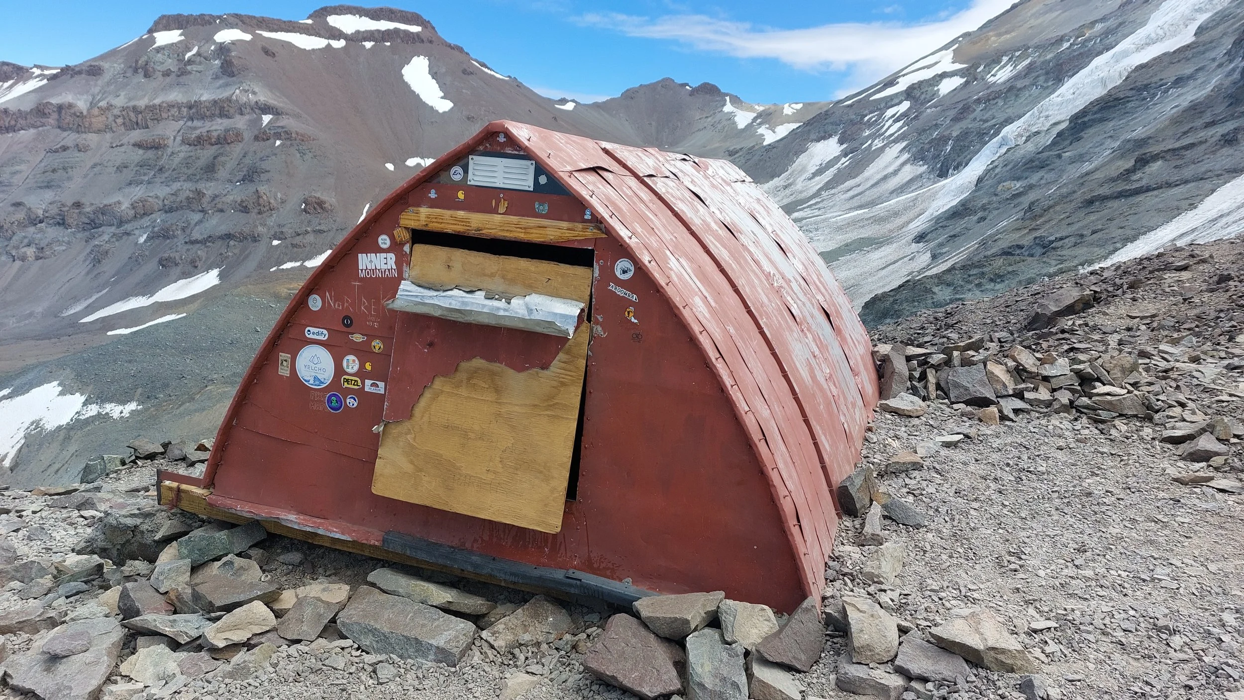 Refugio de montaña rojo en un paisaje de picos nevados y roca, en un entorno de alta montaña.