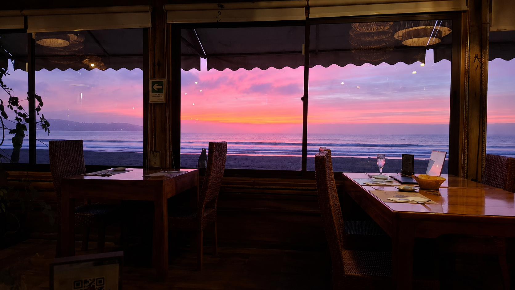 Vista de una playa al atardecer vista desde un restaurante con mesas y sillas de madera frente a la ventana, mostrando un cielo colorido y el mar.