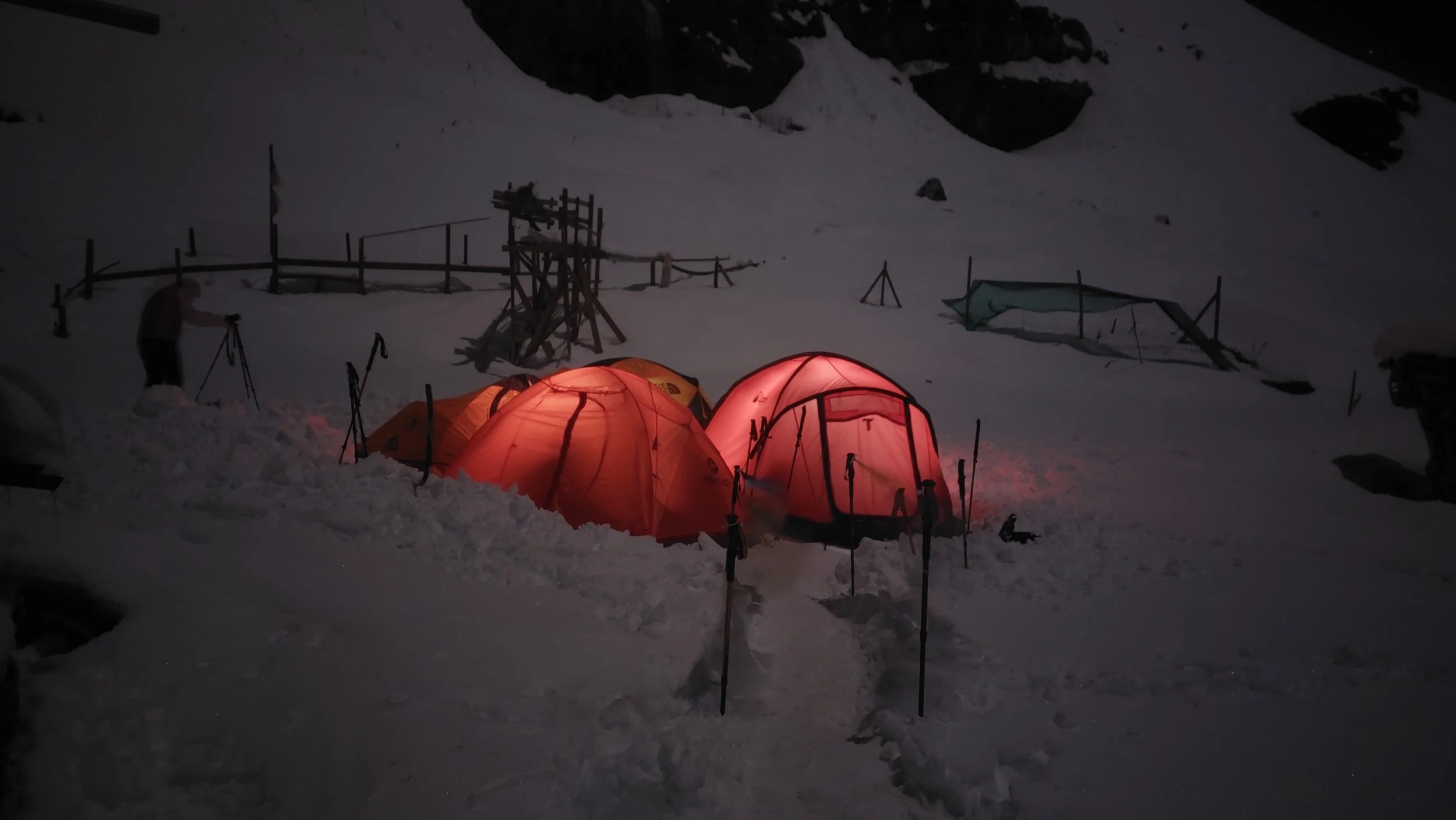 Campamento en la nieve con tiendas de campaña de color naranja iluminadas, en un paisaje montañoso de noche con estructuras de madera y una persona ajustando o preparando equipo.