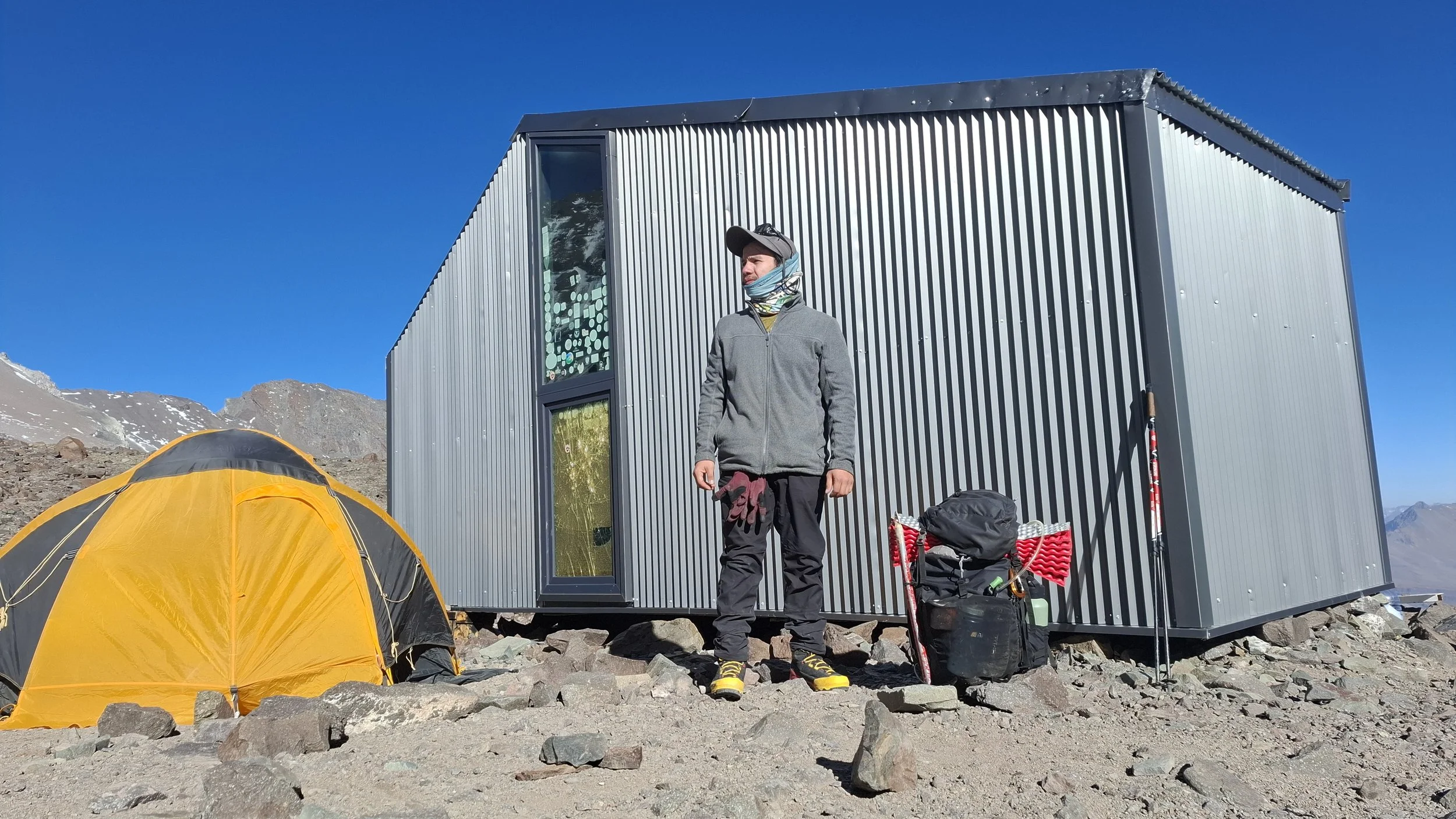 Viajero de montaña con ropa de abrigo, gorra y máscara facial, de pie frente a una estructura metálica en un paisaje de picos nevados y cielo despejado, con una tienda de campaña amarilla y equipo de senderismo alrededor.
