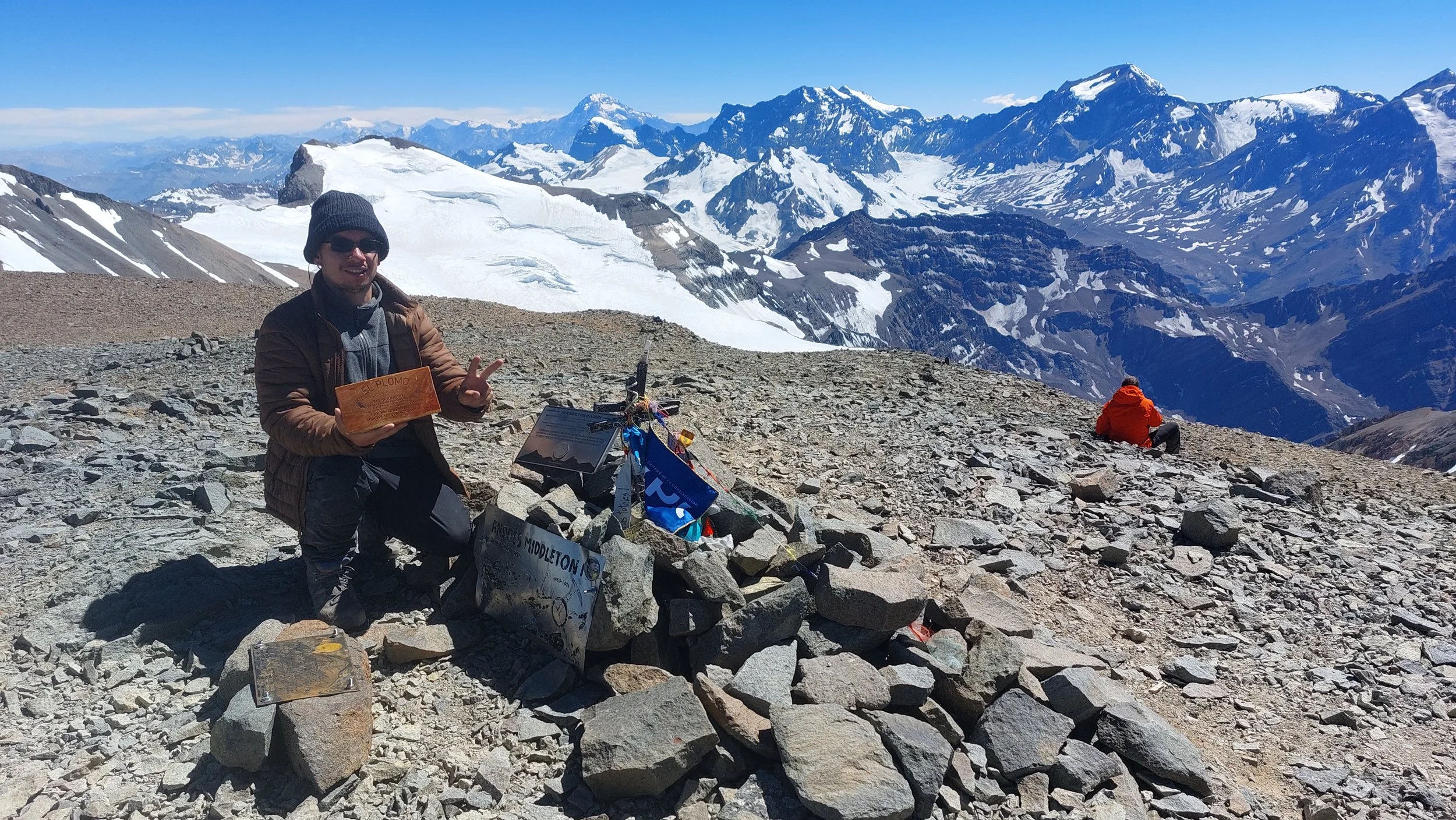 Persona en la cima de una montaña con vista a picos nevados y un cielo azul, sosteniendo un certificado con fondo de montañas y nieve.
