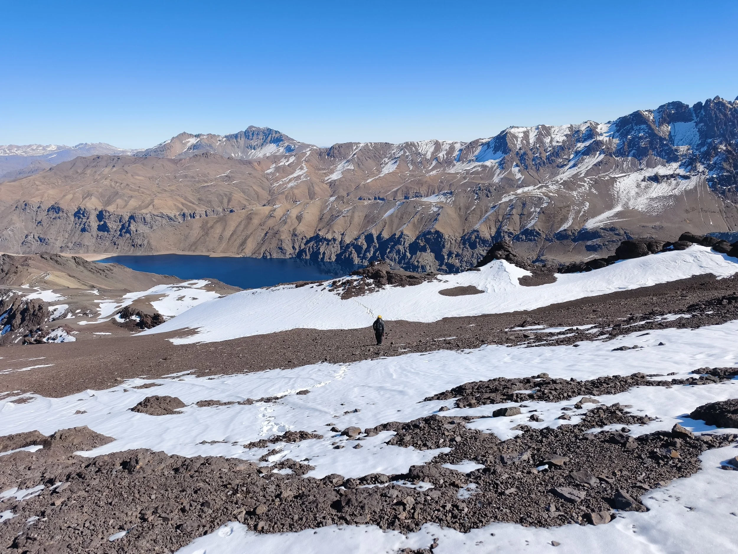Montañas nevadas con picos elevados, un lago en el valle y un caminante con equipo en el primer plano en un entorno de alta montaña.