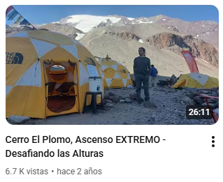 Personas en un campamento con tiendas de campaña en una zona montañosa y rocosa, con montañas y nieve al fondo.