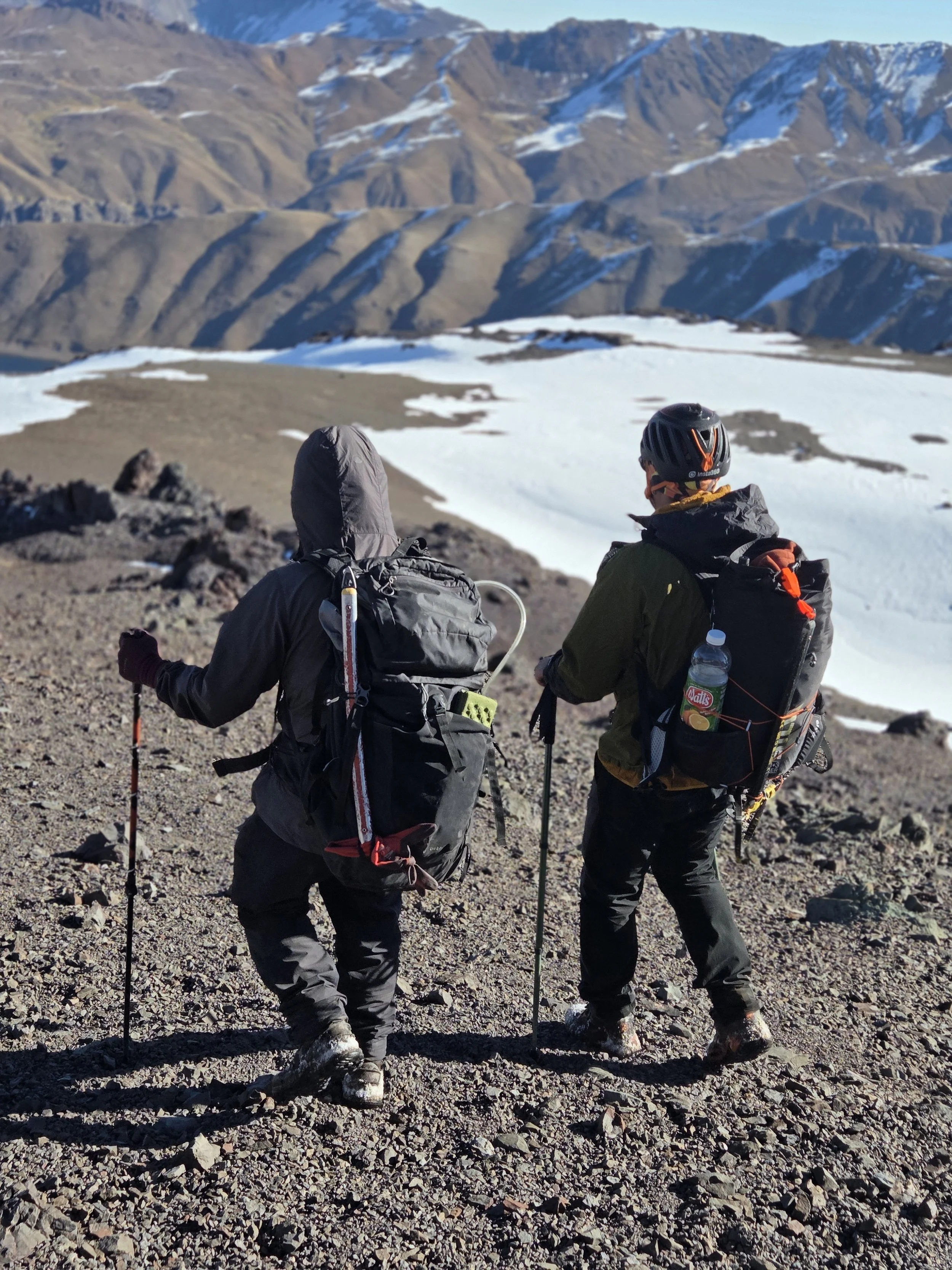 Dos senderistas en paisaje de montaña con nieve y picos escarpados en segundo plano, uno con casco y mochila, el otro con gorro, ambos con bastones de caminata